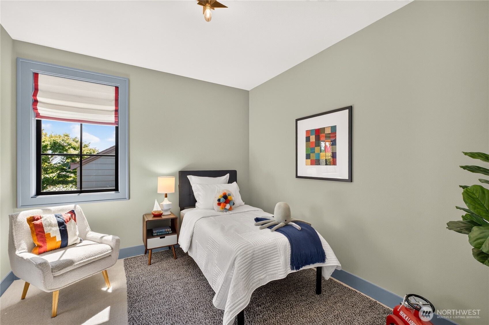 Staged bedroom with a white-covered bed, colorful plush toys, and a white accent chair beside the window.