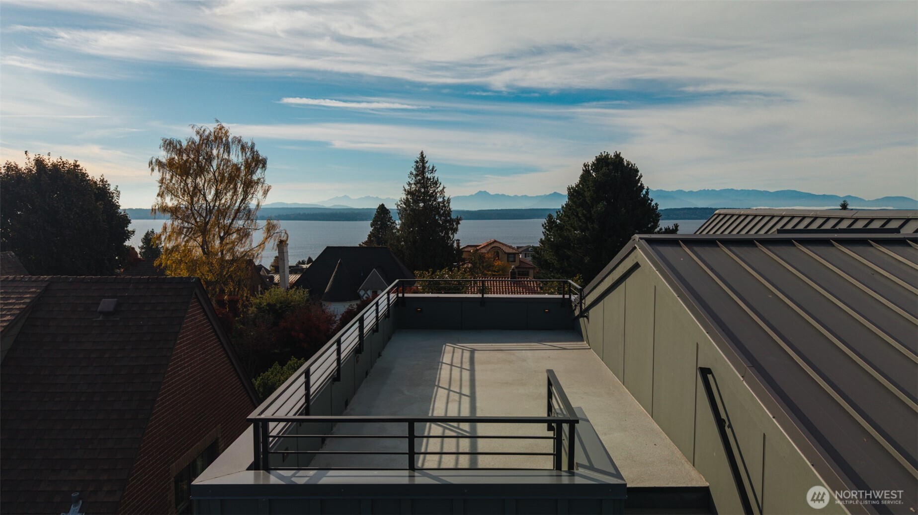 Rooftop terrace overlooking water and distant mountains, with dark railings and neighboring homes.