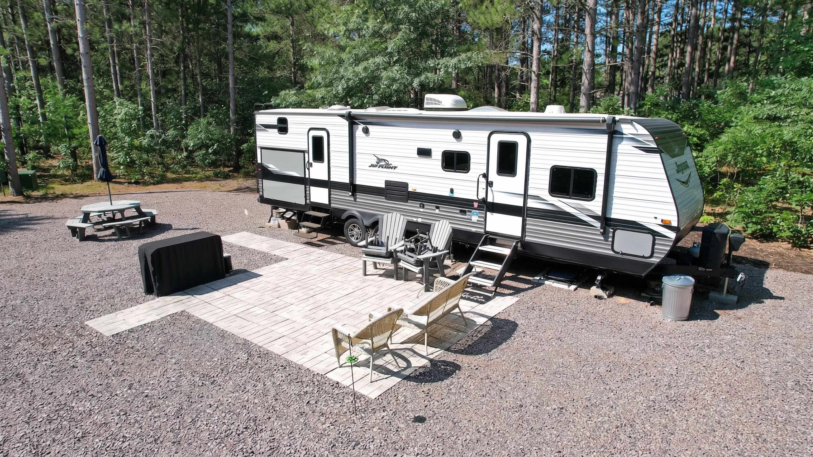 Jay Flight travel trailer at a wooded campsite with a paved patio and outdoor seating.