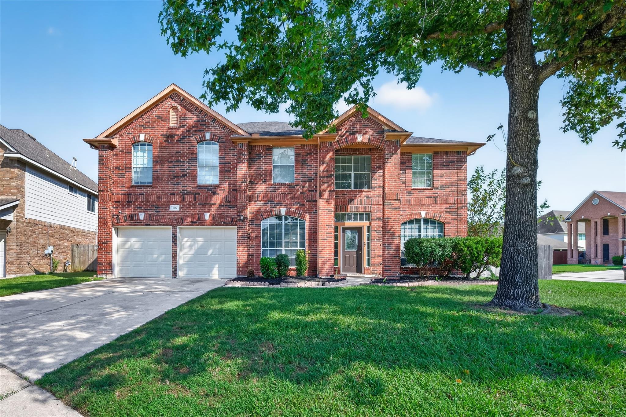 Two-story red brick house with double garage doors, arched windows, and a large shade tree in front.