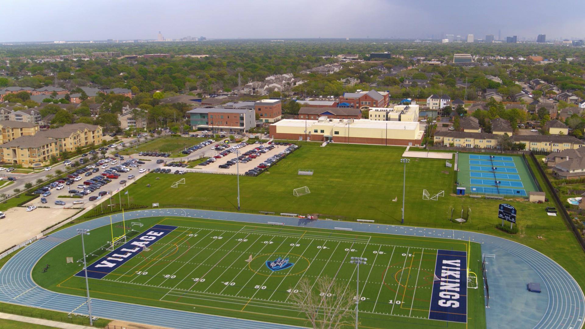 Aerial view of a football field, blue running track, practice fields, and tennis courts, with Houston’s skyline in the distance.