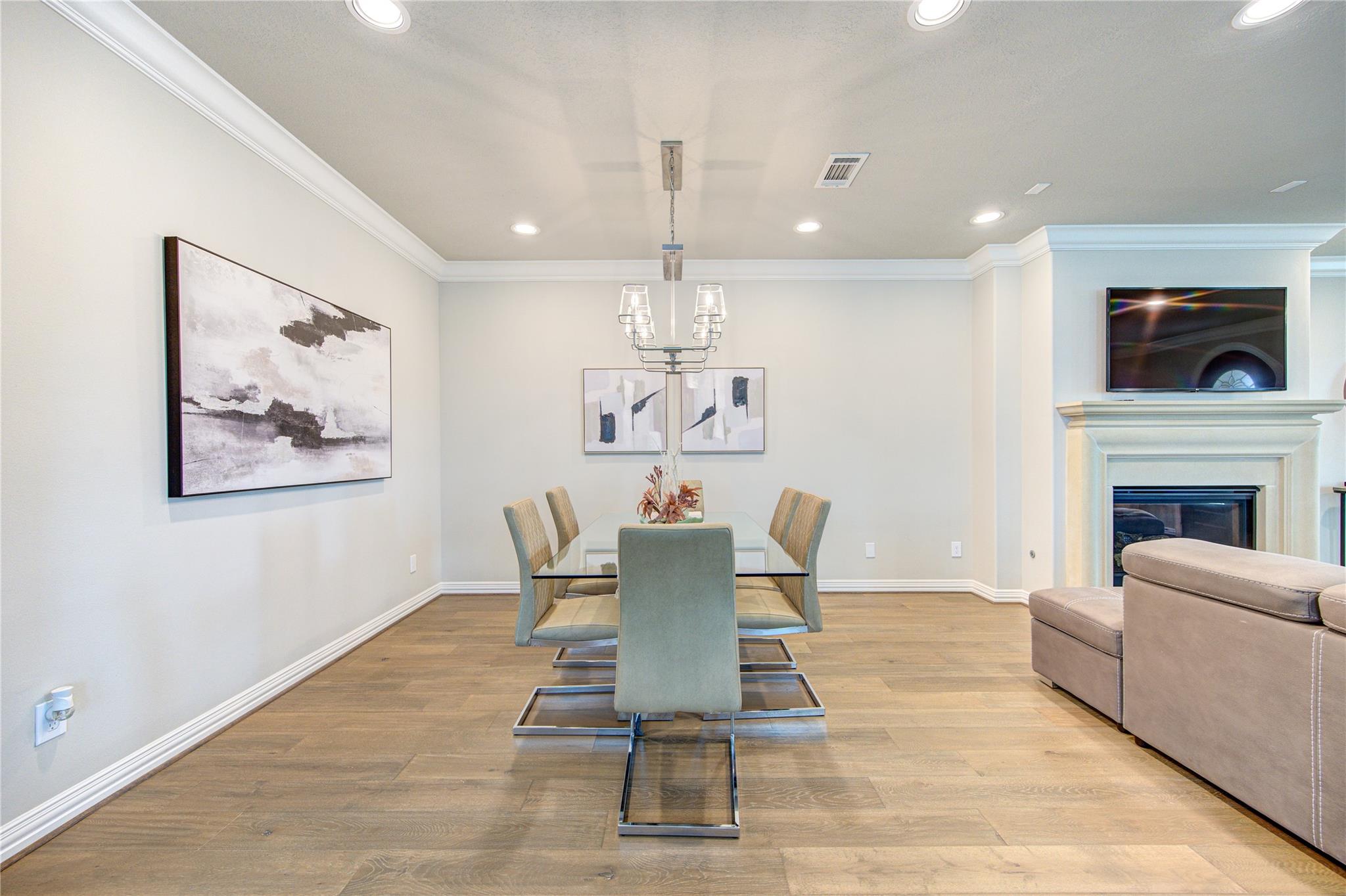 Dining room with a glass table, upholstered chairs, and chandelier beside a living area fireplace.