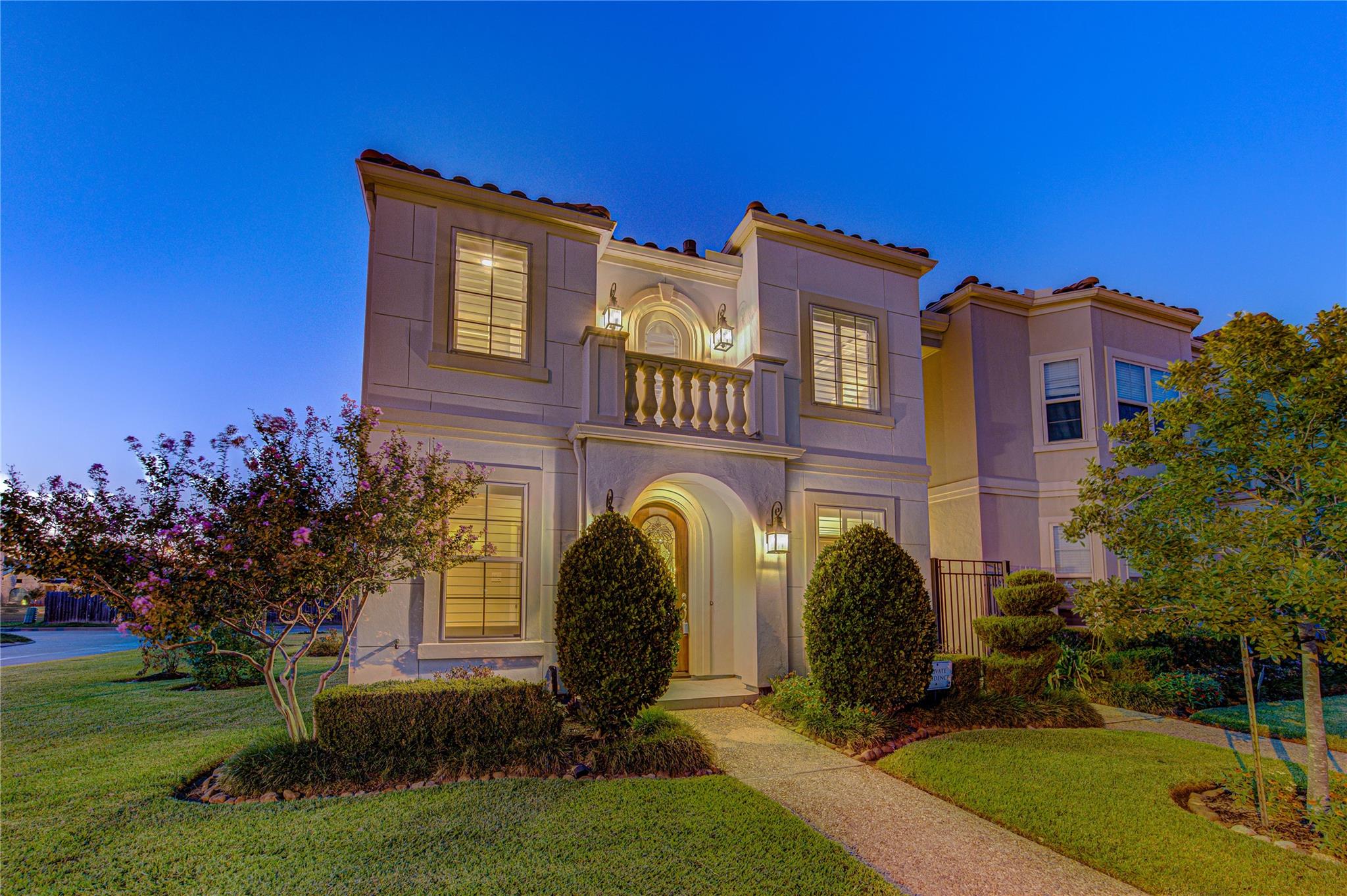 Two-story stucco home with an arched entry, balcony, and landscaped front yard at dusk.