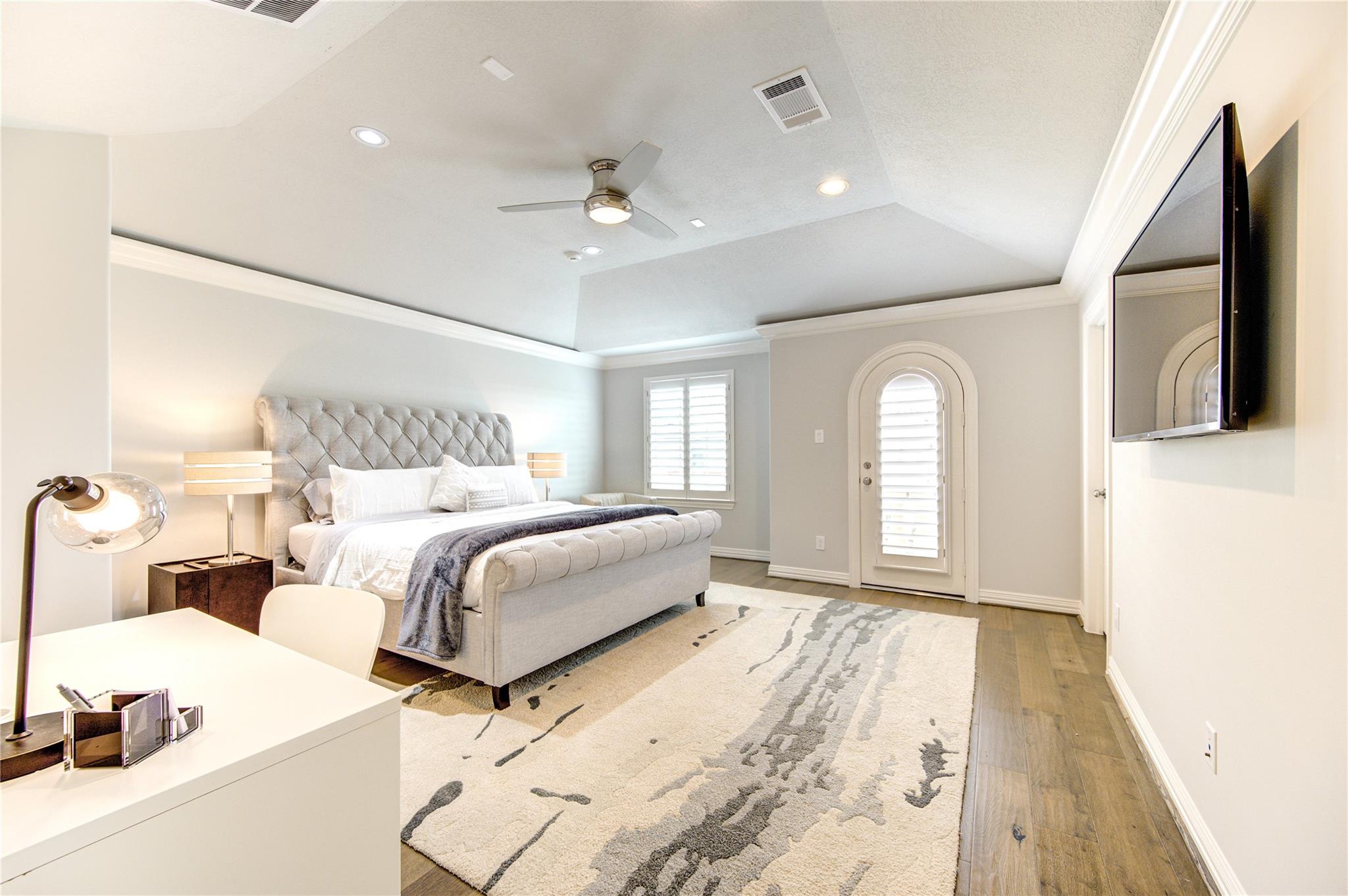 Furnished bedroom with a tufted bed, patterned rug, tray ceiling, and arched shuttered door.