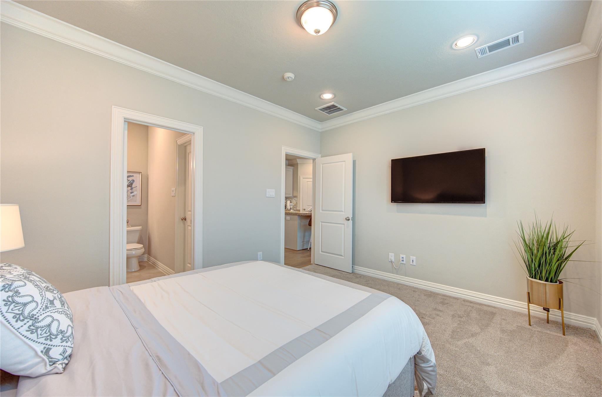 Staged bedroom with a white bed, wall-mounted television, and open doorway to the bathroom.