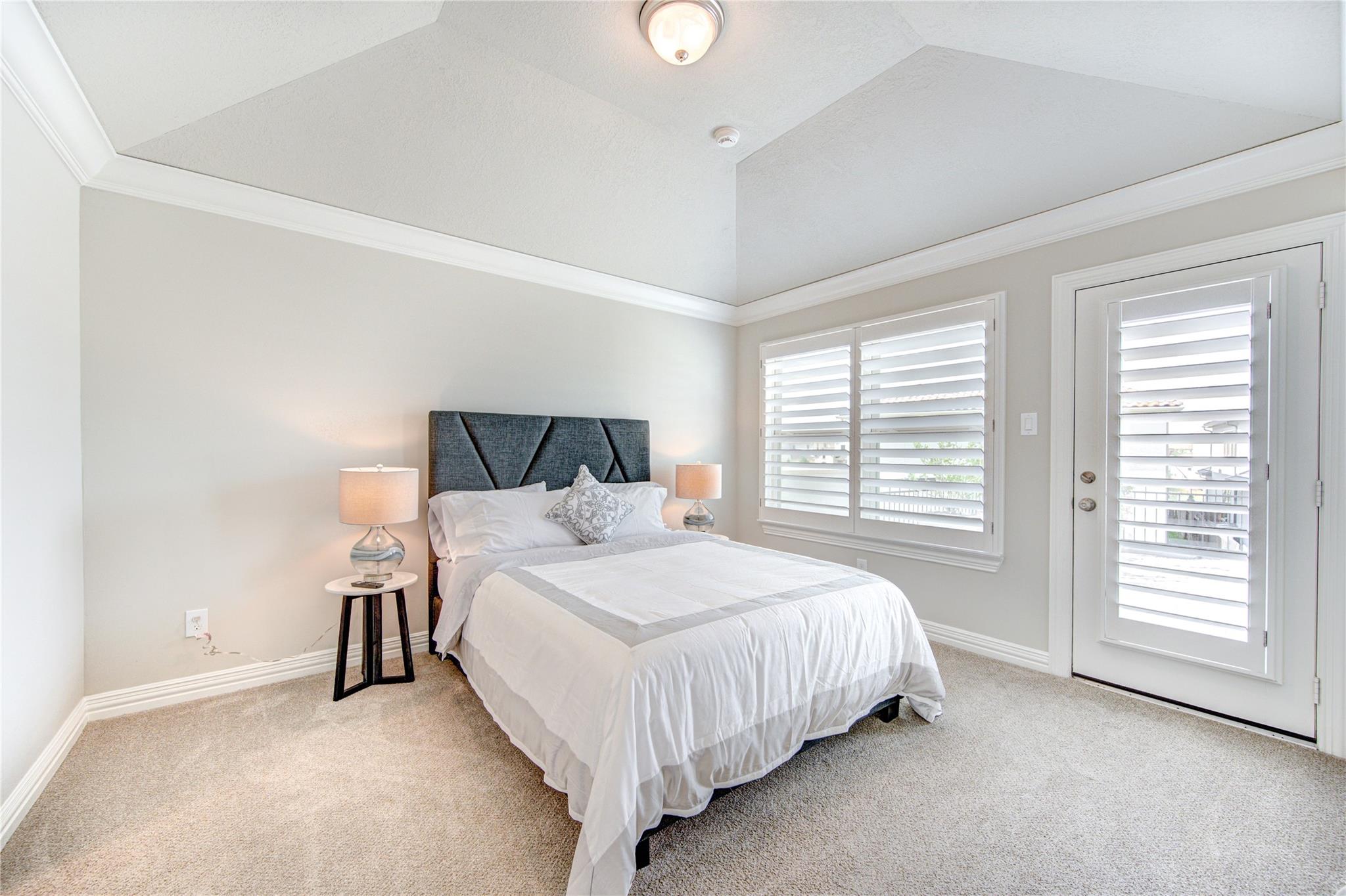 Staged bedroom with a white bed, dark upholstered headboard, tray ceiling, and shuttered windows and door.