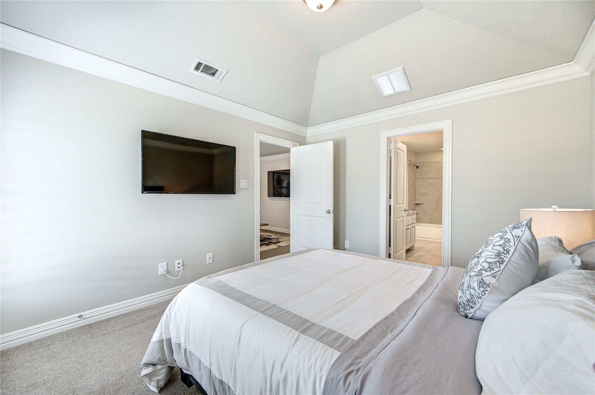 Bedroom with a white and gray bed, wall-mounted TV, vaulted ceiling, and adjoining bathroom doorway.