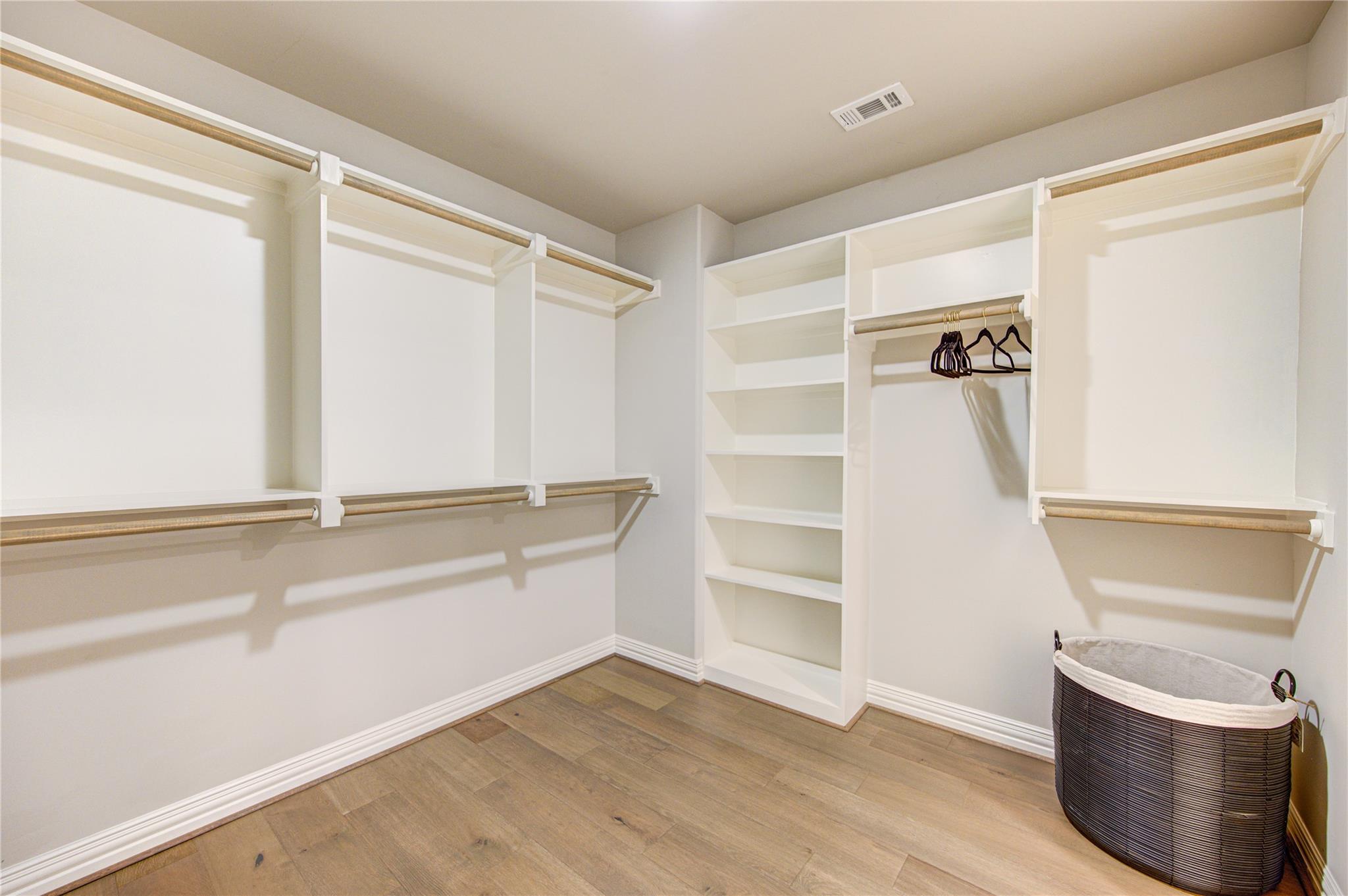 Walk-in closet with white built-in shelves and hanging rods, wood flooring, and a woven hamper.