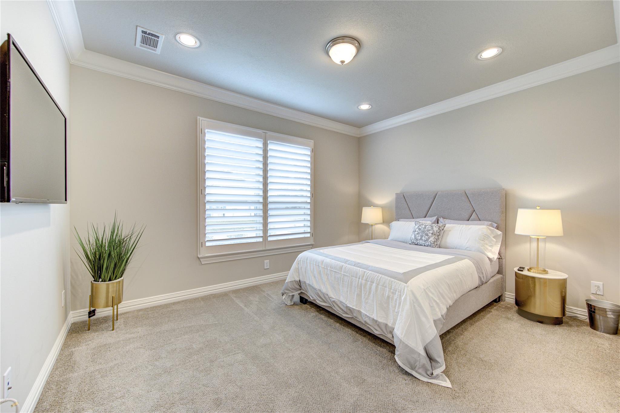 Staged bedroom with a gray upholstered bed, white bedding, plantation shutters, and beige carpet.