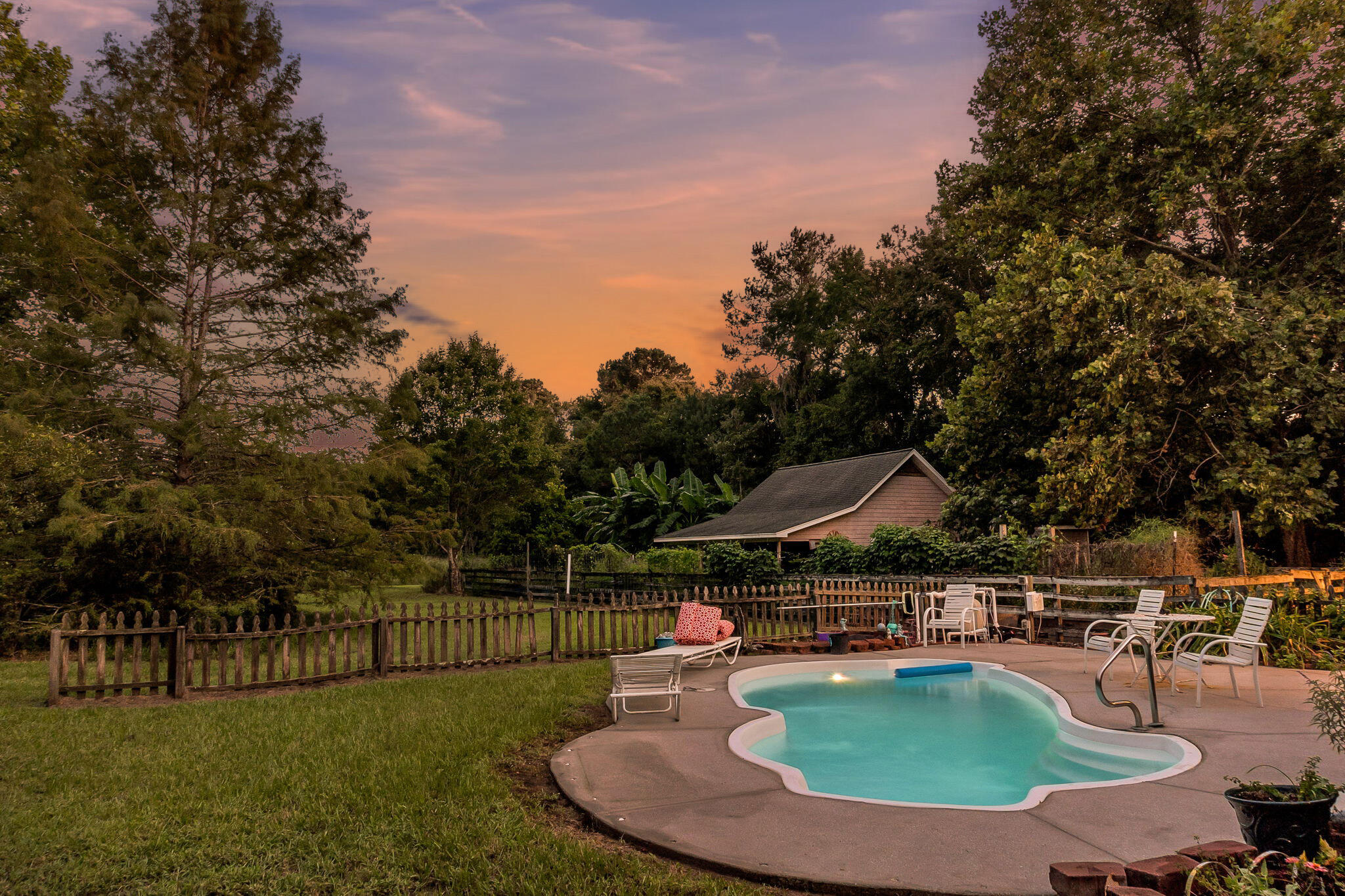 Backyard pool and patio with white lounge chairs, a wooden fence, and mature trees at dusk.