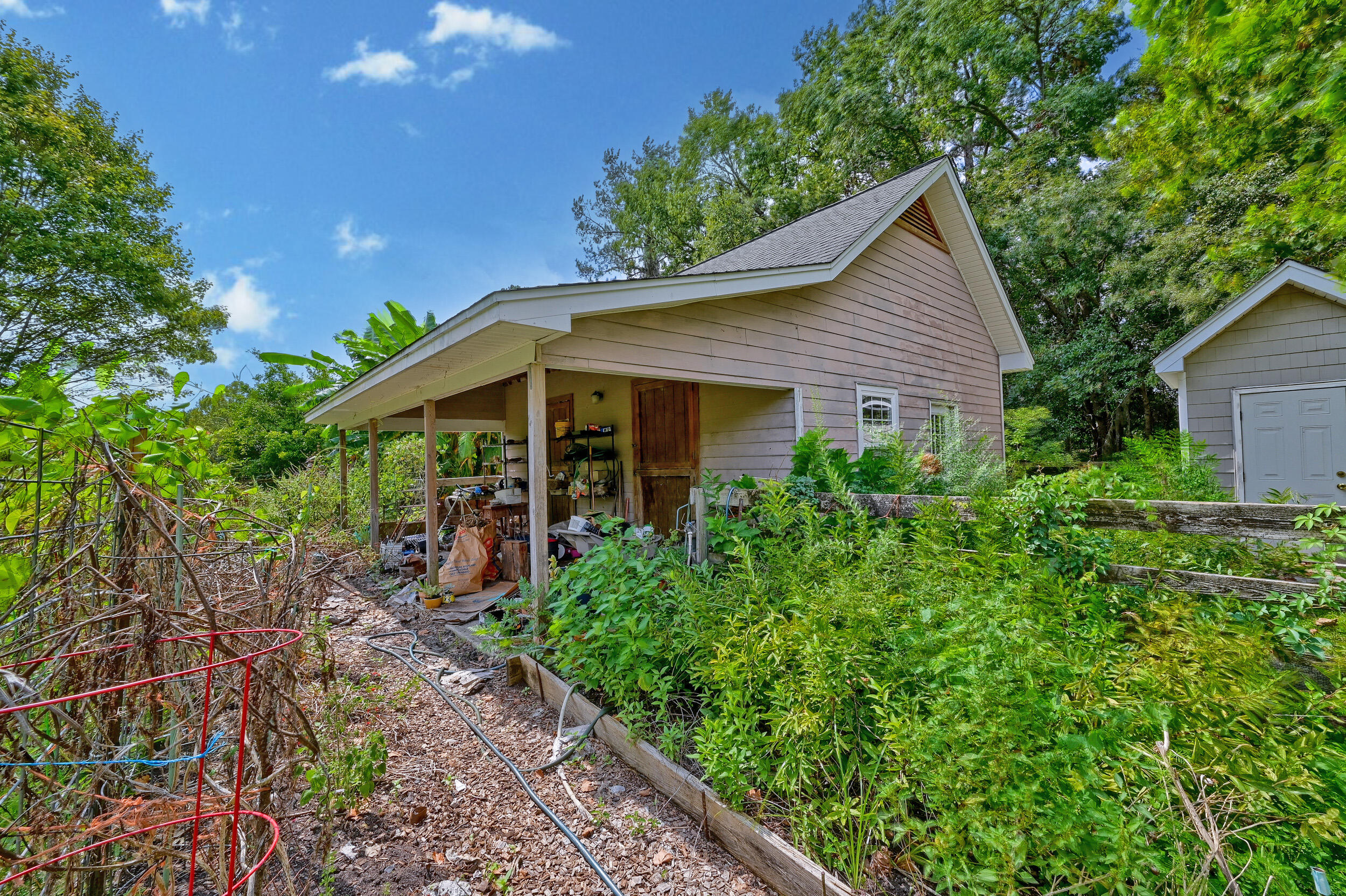 Taupe-sided house with a cluttered covered work area amid dense greenery.