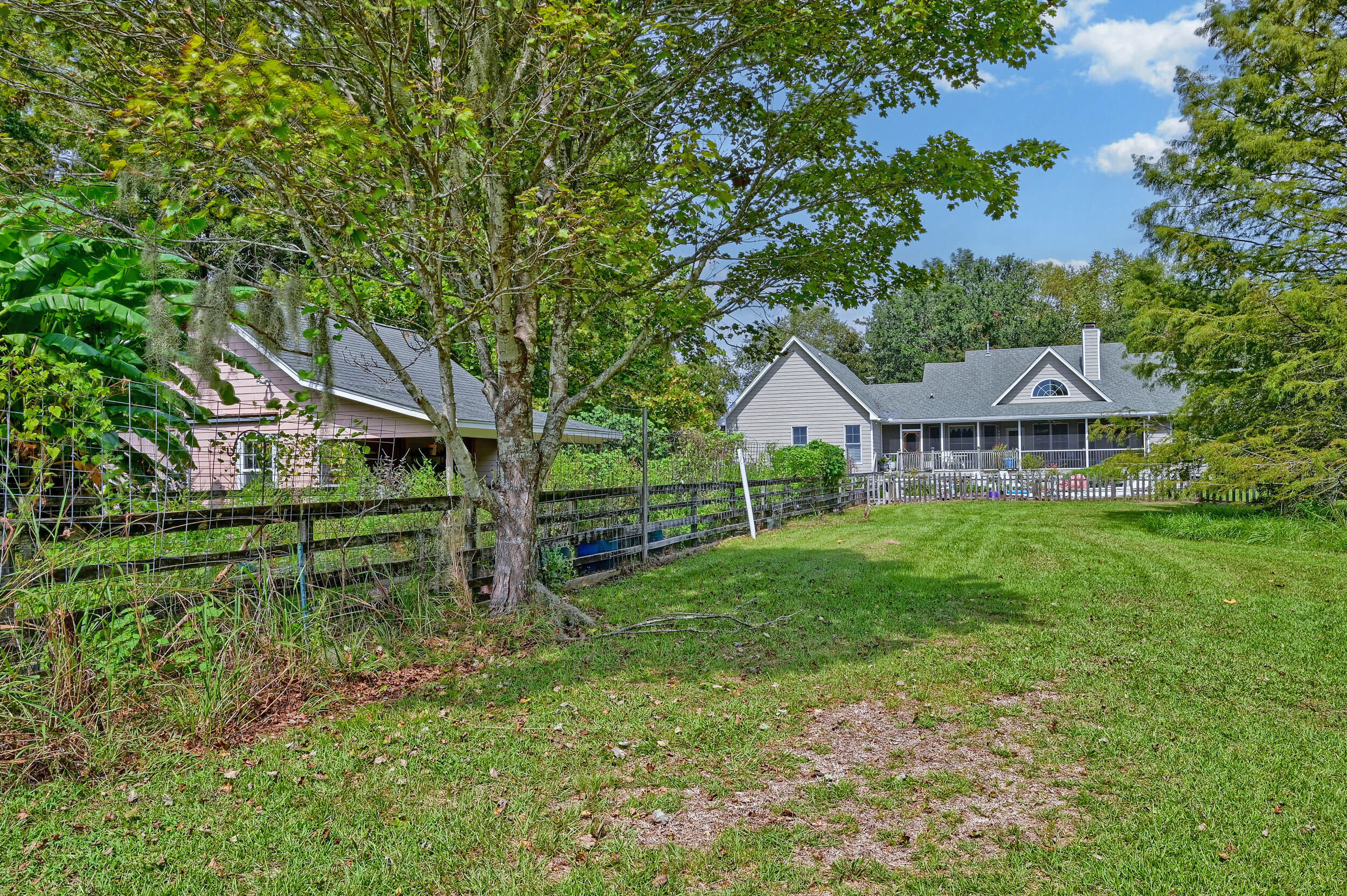 Rear of a light-colored home beyond a broad lawn, with a covered porch, fenced garden, and mature trees.