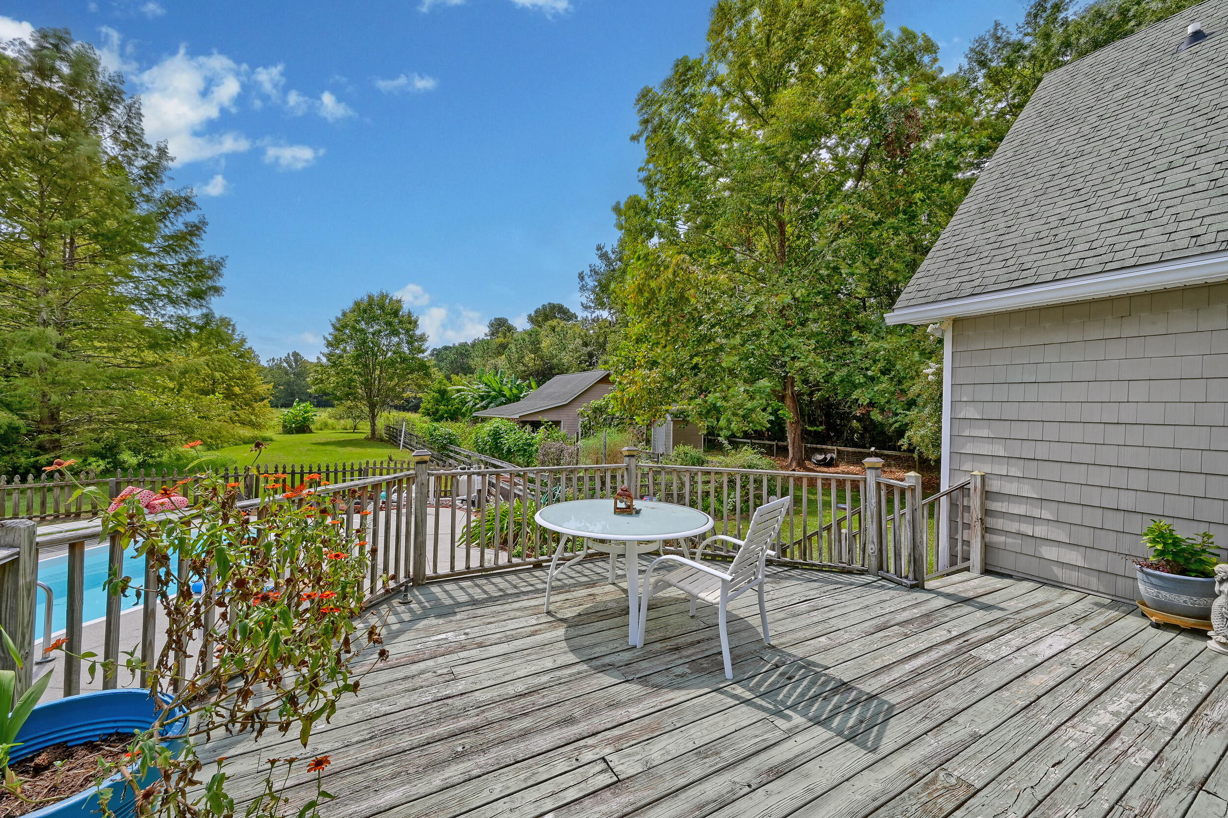 Wooden deck with white patio furniture overlooking a pool, flower beds, and a leafy yard.