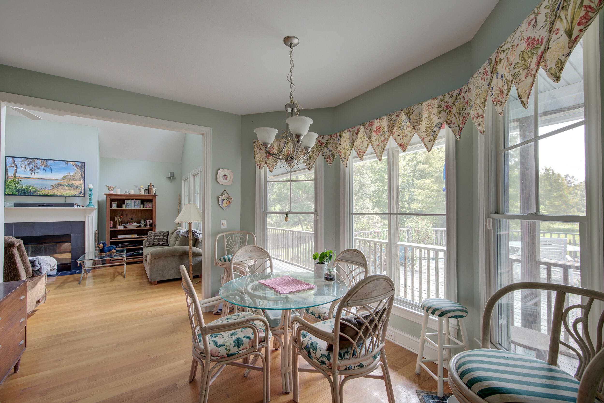 Dining area with glass table and wicker chairs beside tall windows overlooking a deck and trees.