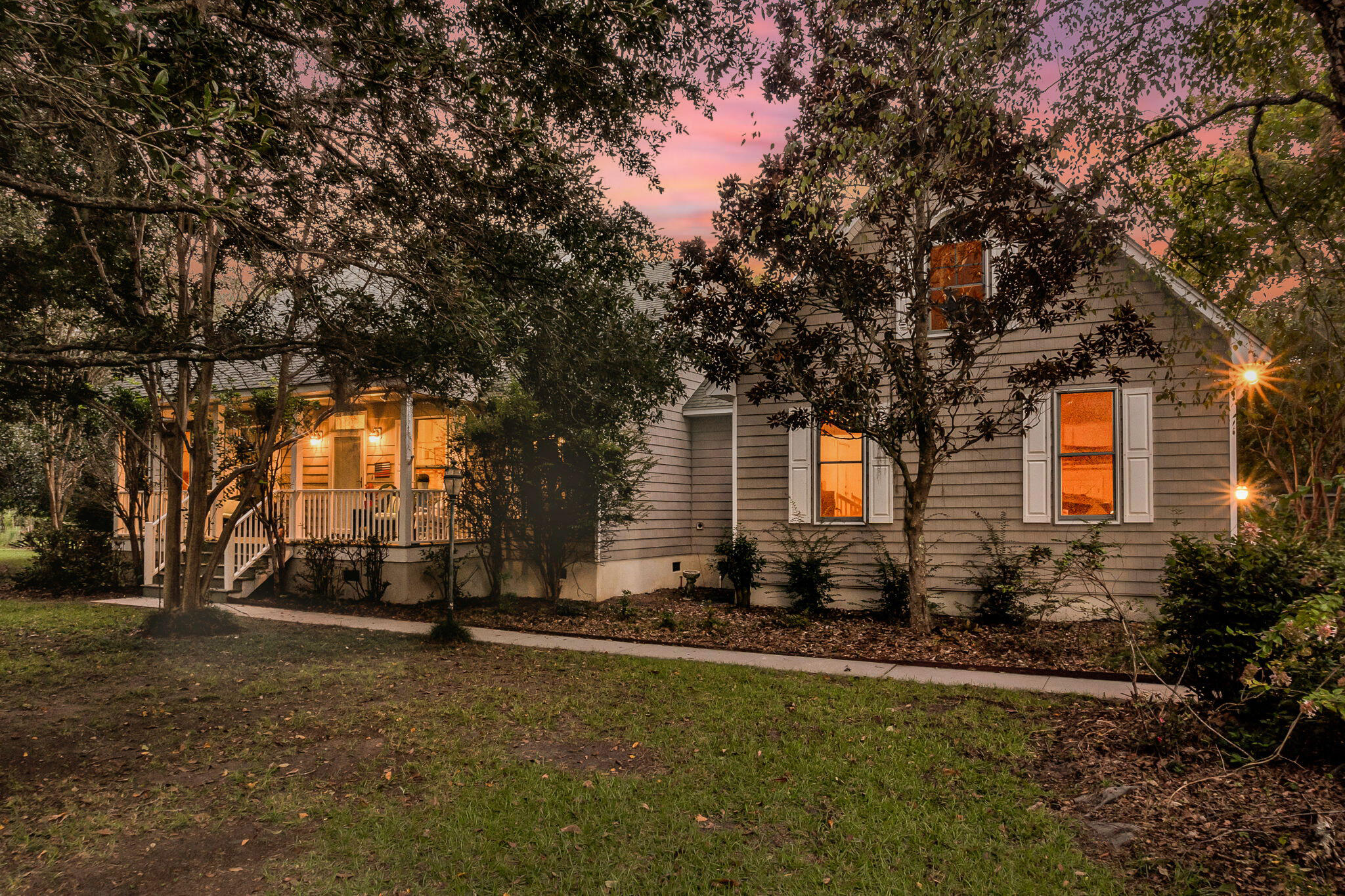 Gray-sided house with a covered porch and glowing windows framed by trees at dusk.