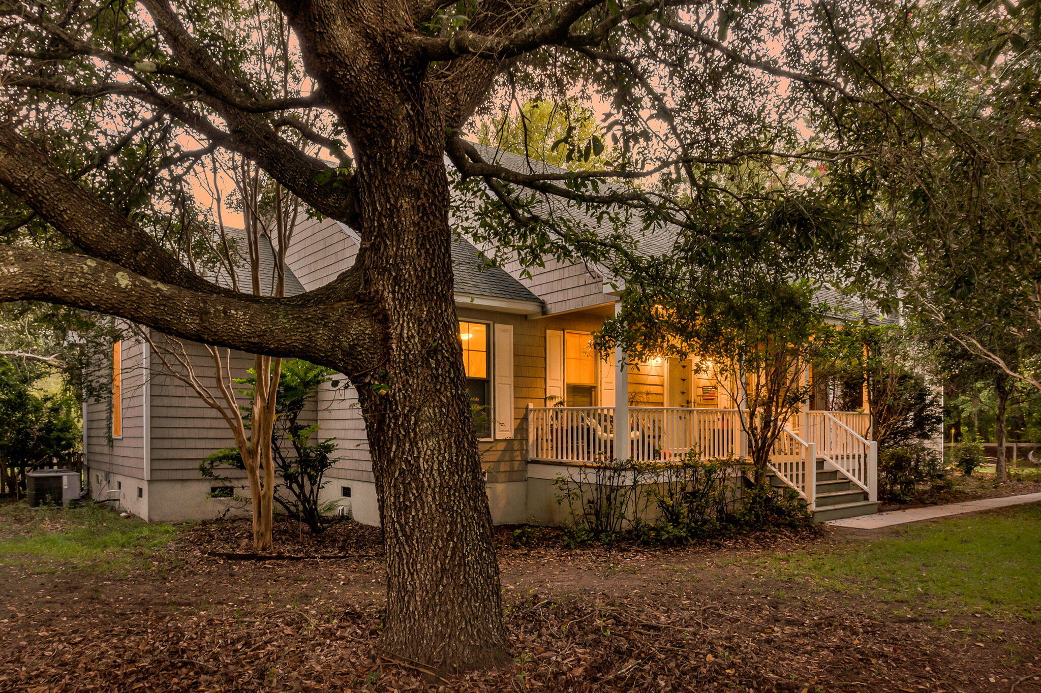 Gray-sided home with a warmly lit front porch, framed by a large oak tree at dusk.