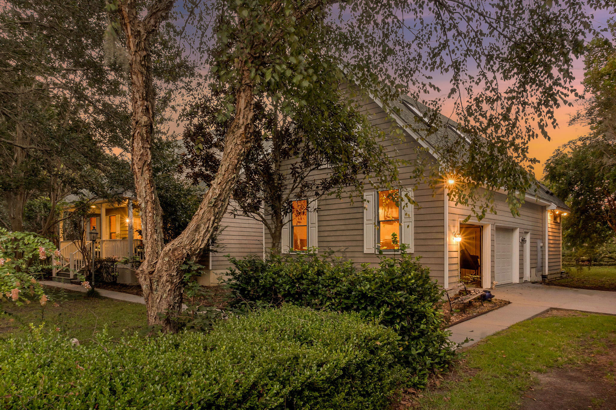 Gray clapboard house with a covered porch and attached garage, framed by mature trees at sunset.