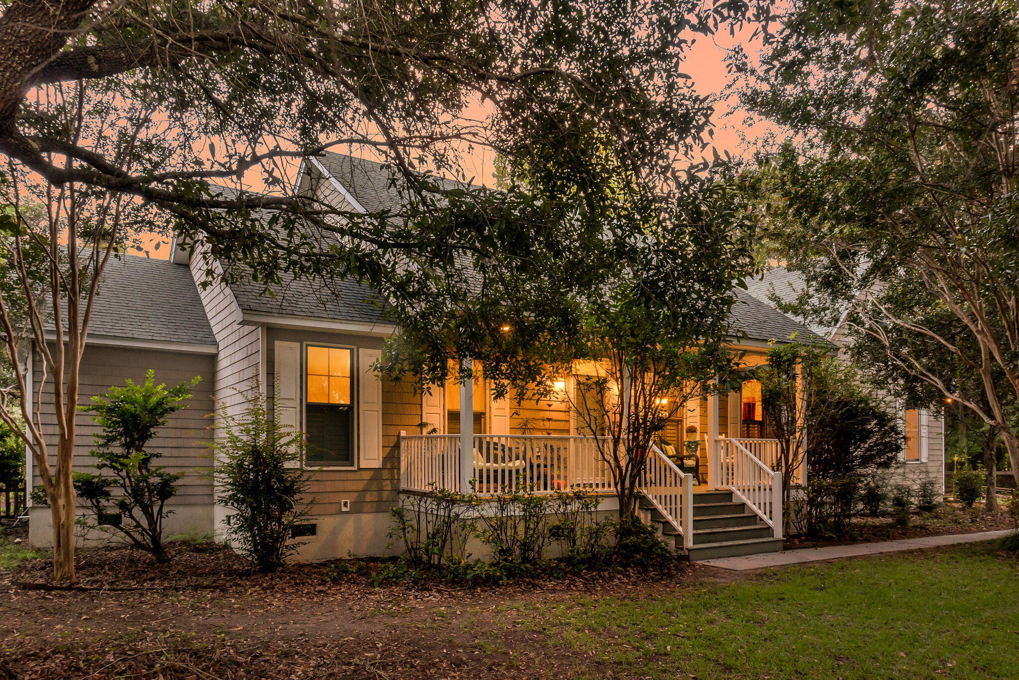 Gray-shingled house with a warmly lit covered porch and white railings, framed by mature trees at dusk.
