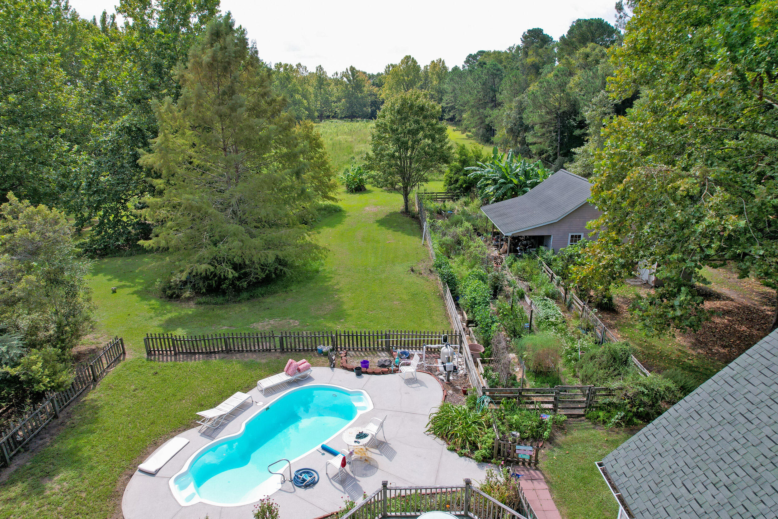 Backyard with a swimming pool and patio, expansive lawn, fenced garden beds, and surrounding mature trees.