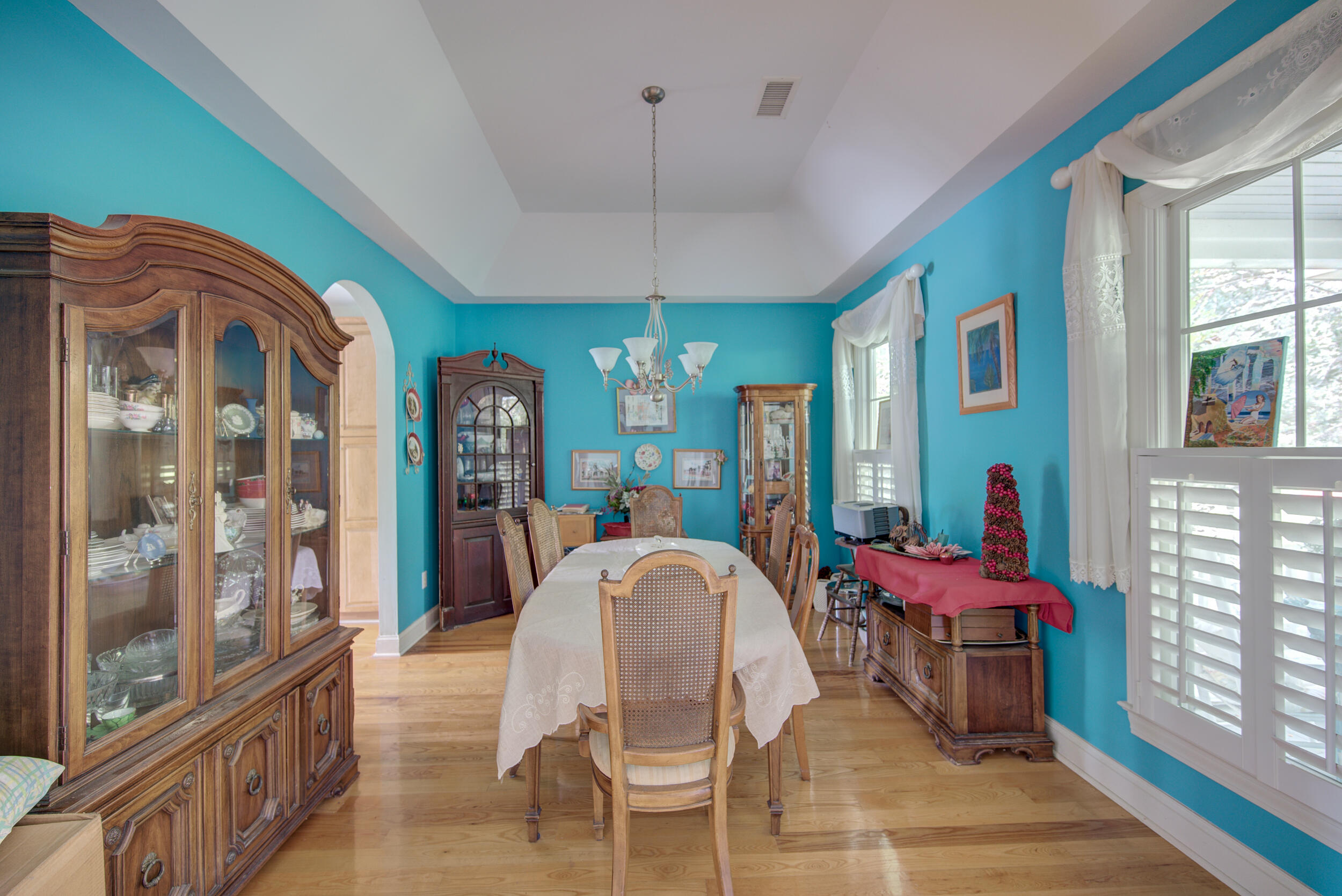 Dining room with turquoise walls, a long white-covered table, ornate wooden cabinets, and bright windows.