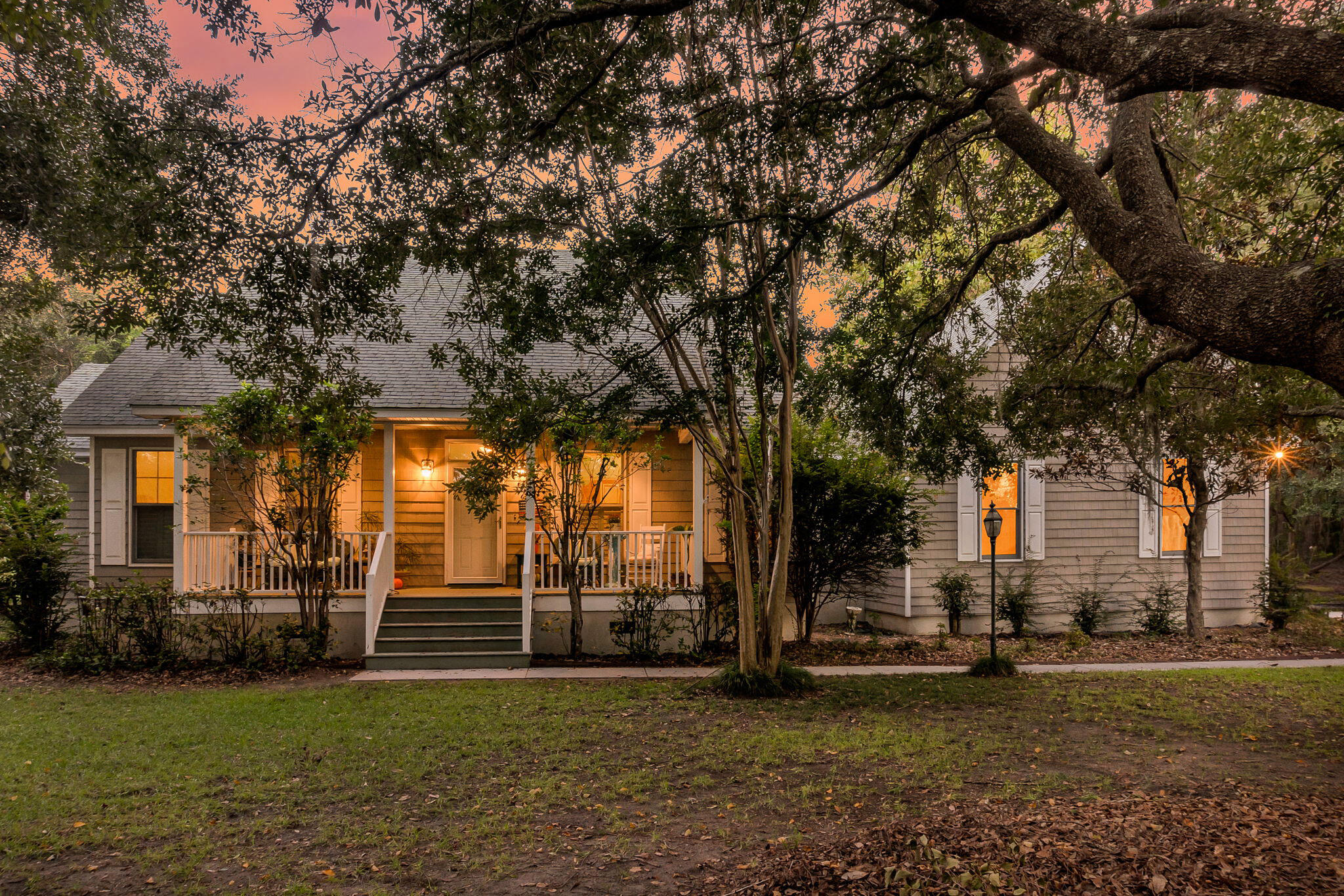 Gray shingle-sided house with a white-railed porch and warmly lit windows beneath mature trees at dusk.