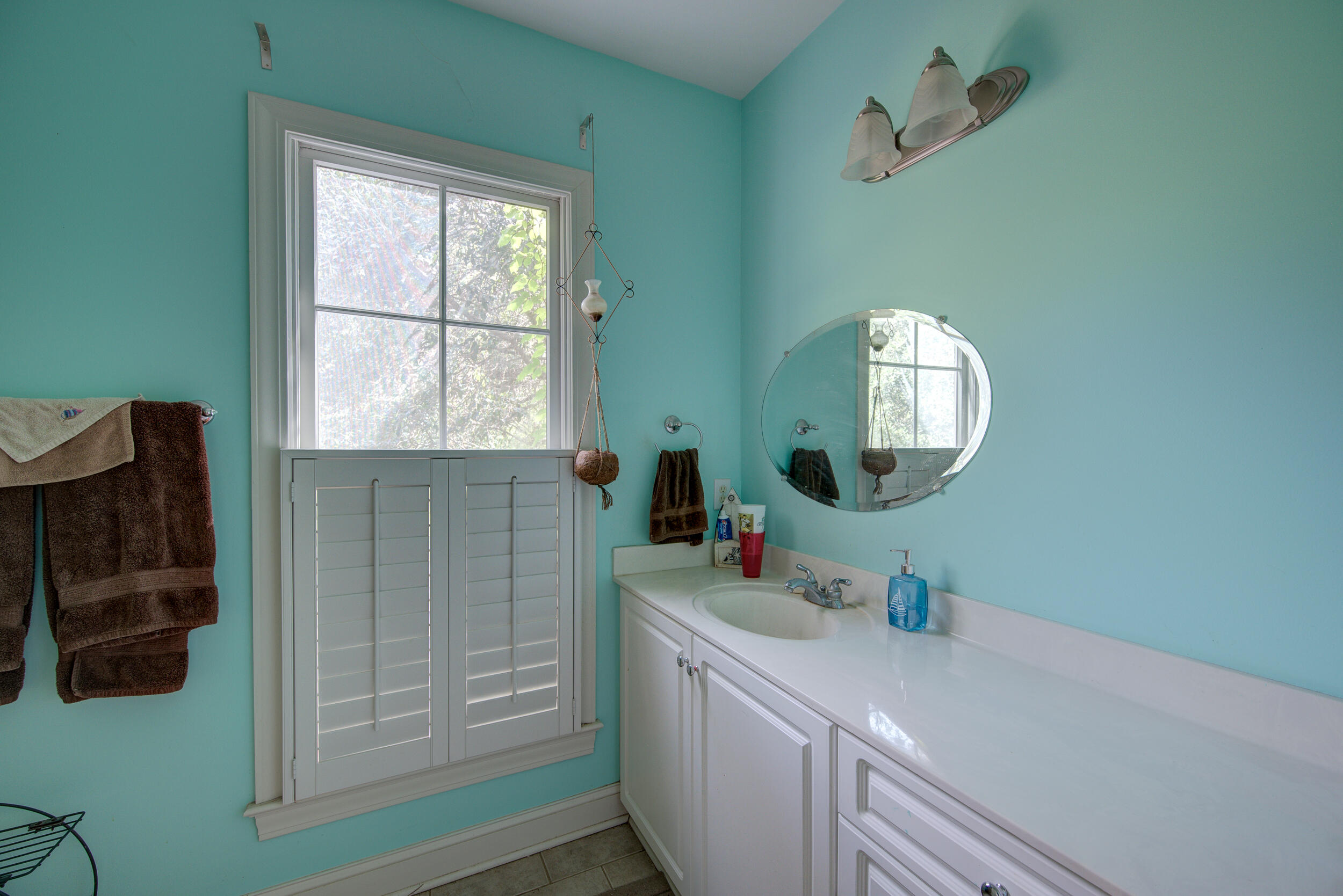 Bathroom with a white vanity, oval mirror, and shuttered window against turquoise walls.