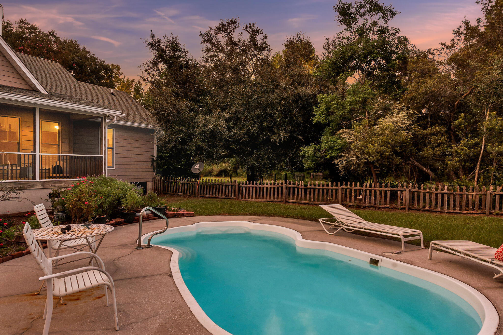 Backyard swimming pool beside a screened porch, patio seating, and wooded fence line at dusk.