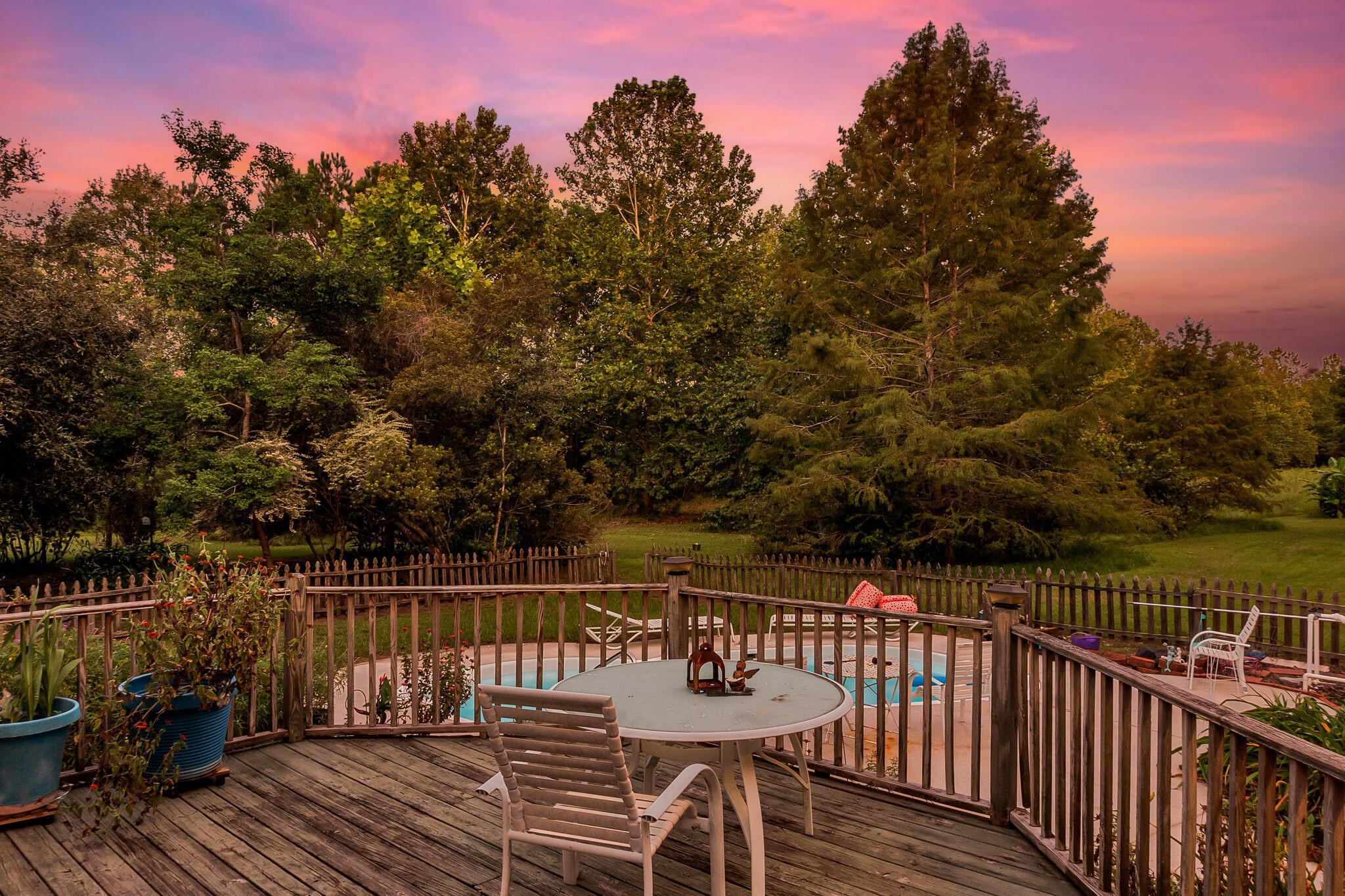 Wooden deck with outdoor dining set overlooking a fenced pool and leafy yard at dusk.
