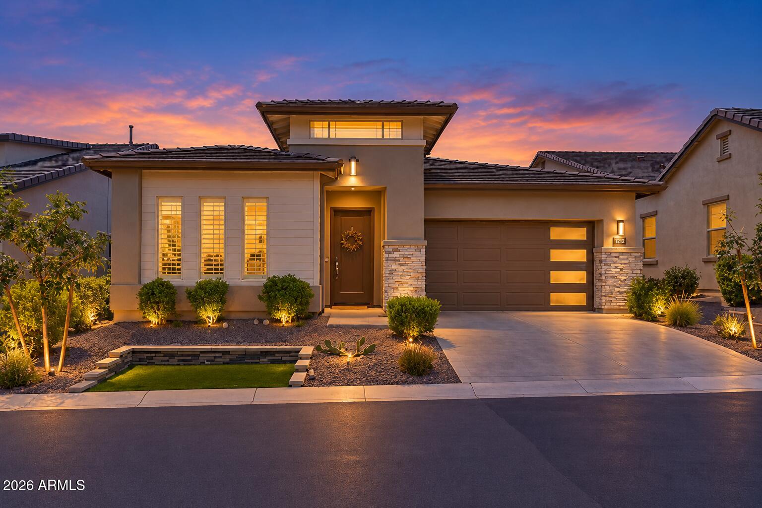Modern stucco home at dusk with illuminated windows, a two-car garage, and desert landscaping.