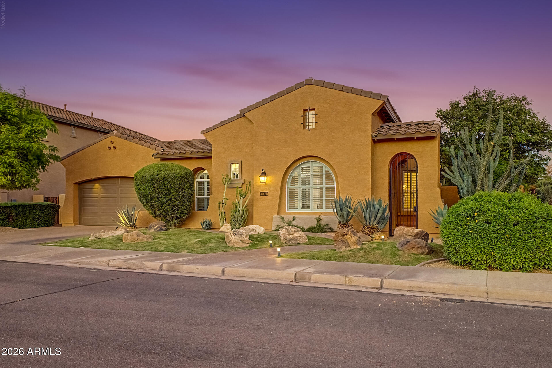 Tan stucco home with arched windows, tiled roof, attached garage, and desert landscaping at dusk.