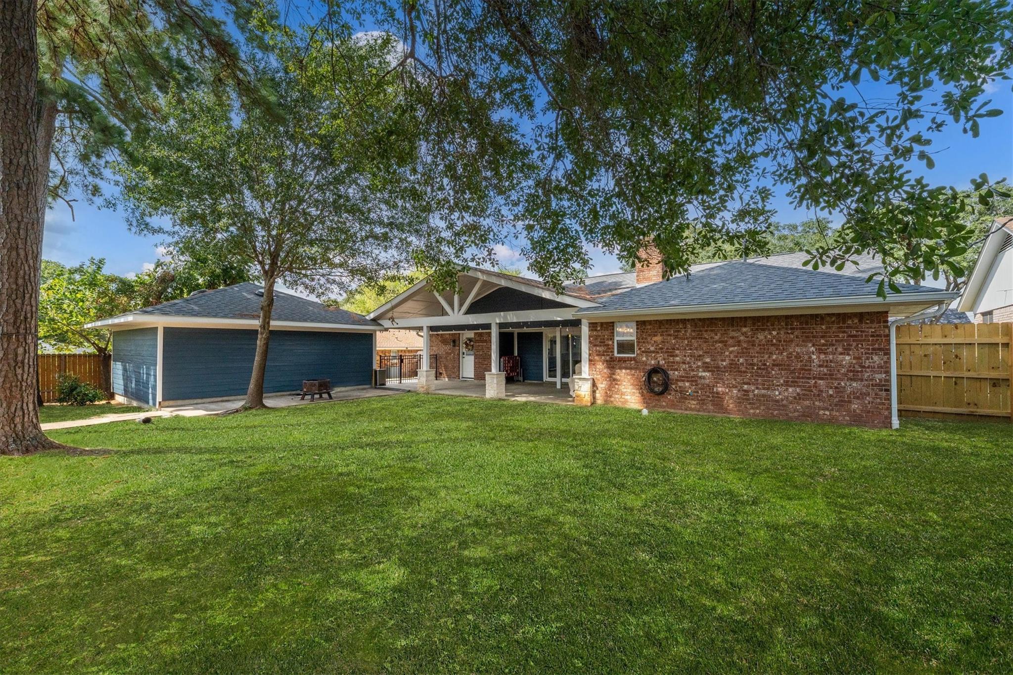 Rear of brick home with covered patio, broad lawn, and mature shade trees.