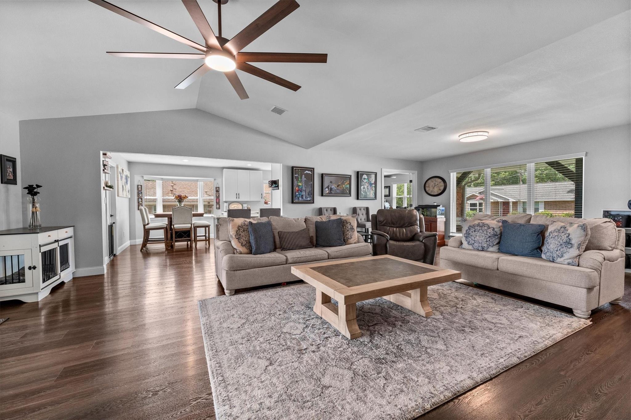 Furnished living room with vaulted ceiling, large ceiling fan, dark wood floors, and adjoining dining area.