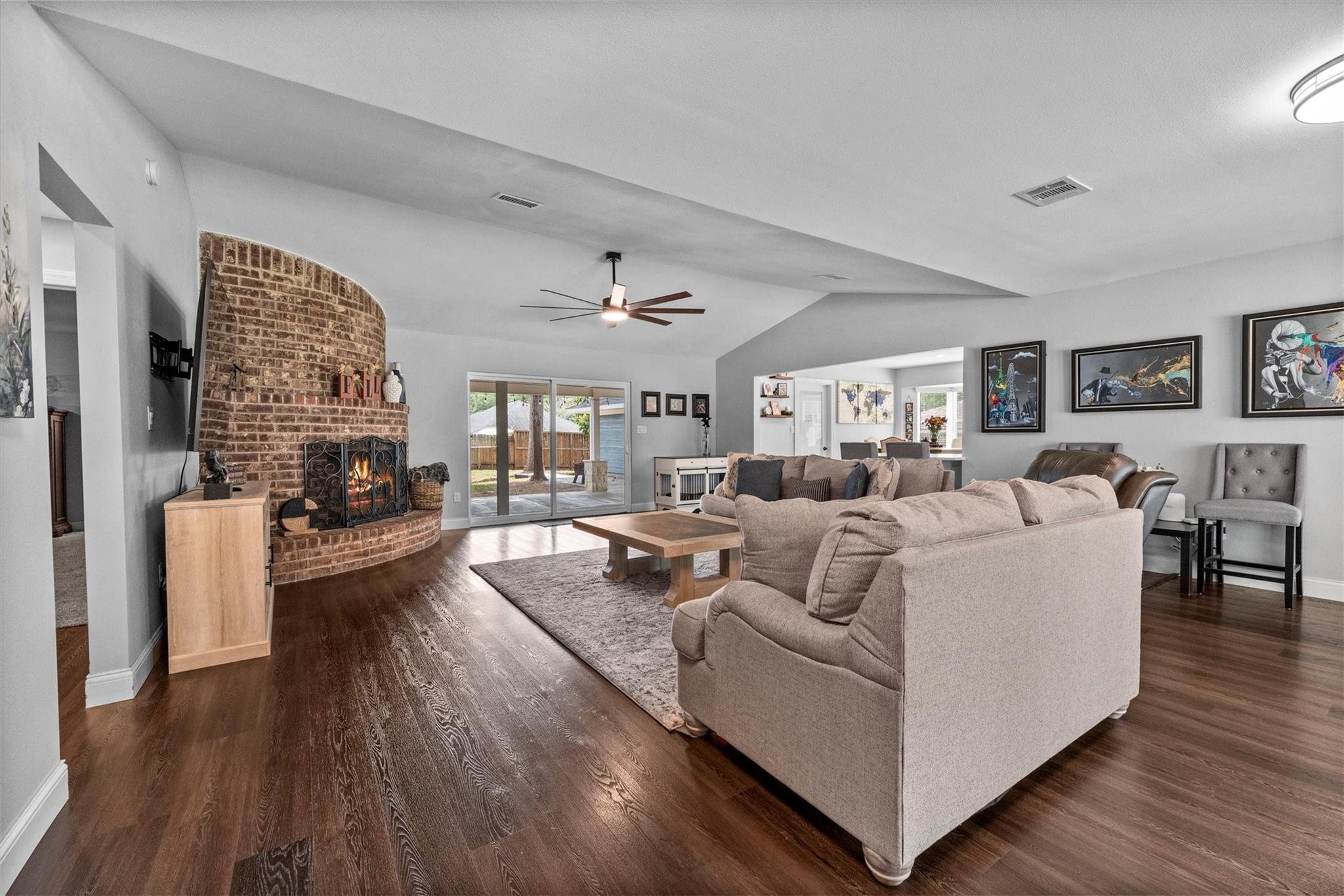 Open living room with a curved brick fireplace, dark wood floors, and sliding glass doors to the backyard.