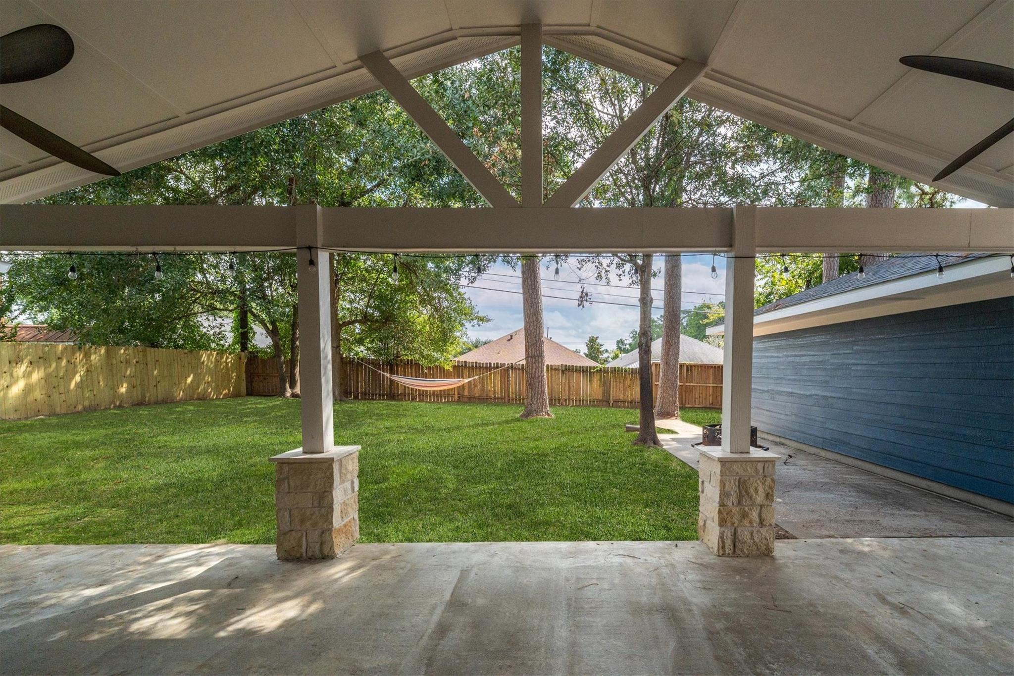 Covered backyard patio overlooking a fenced lawn with mature trees and a hammock.