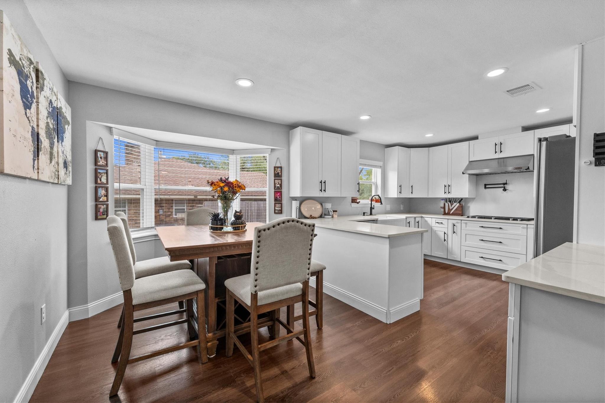 Bright kitchen with white cabinetry and island, wood floors, and a dining table beside a wide window.