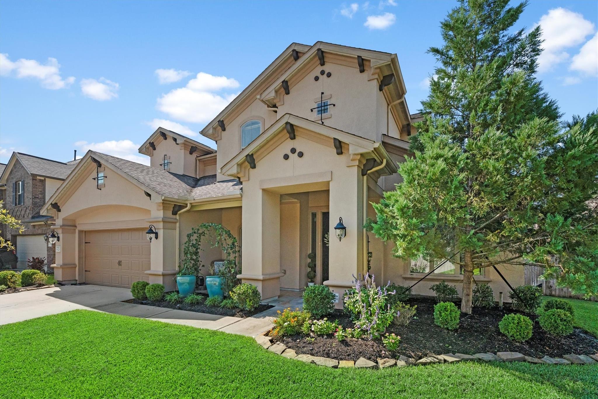 Two-story beige stucco home with a gabled roof, landscaped entry, and large evergreen tree.
