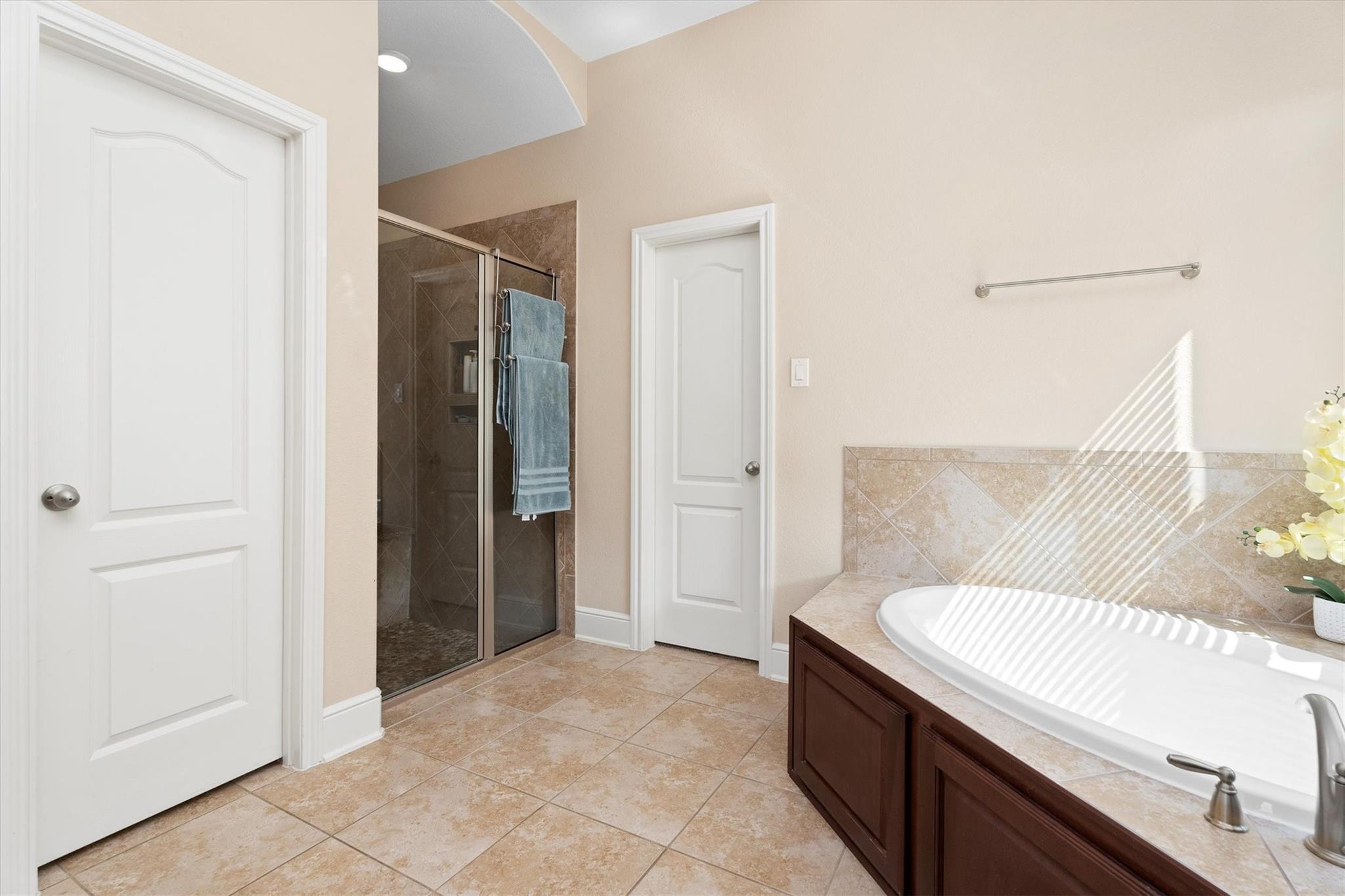 Bathroom with a large soaking tub and glass-enclosed tiled shower; sunlight stripes the tub.