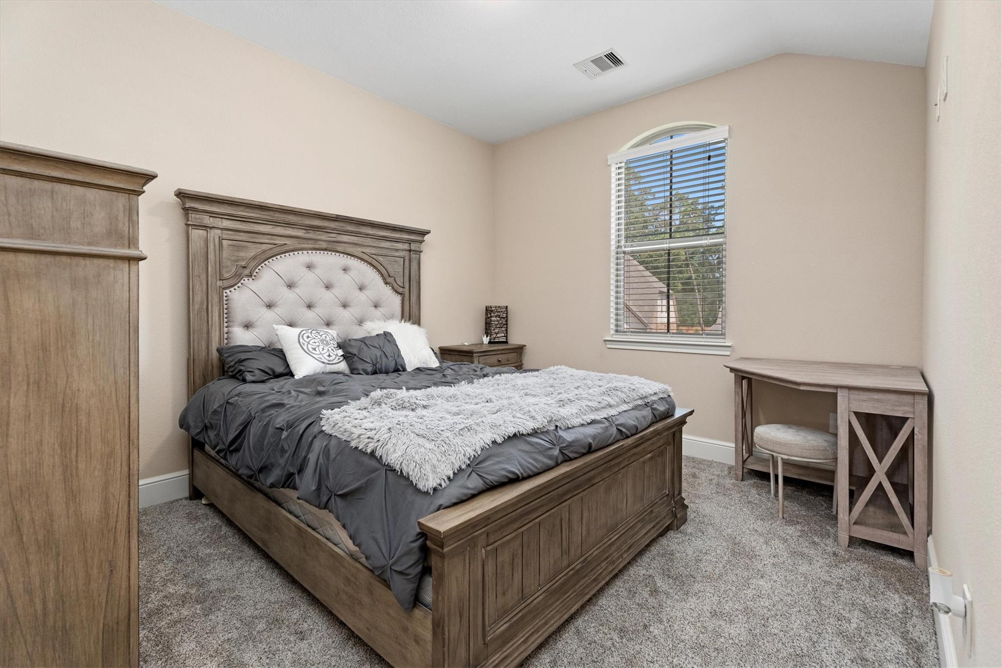 Furnished bedroom with a dark wood bed, tufted headboard, gray bedding, and a window with blinds.