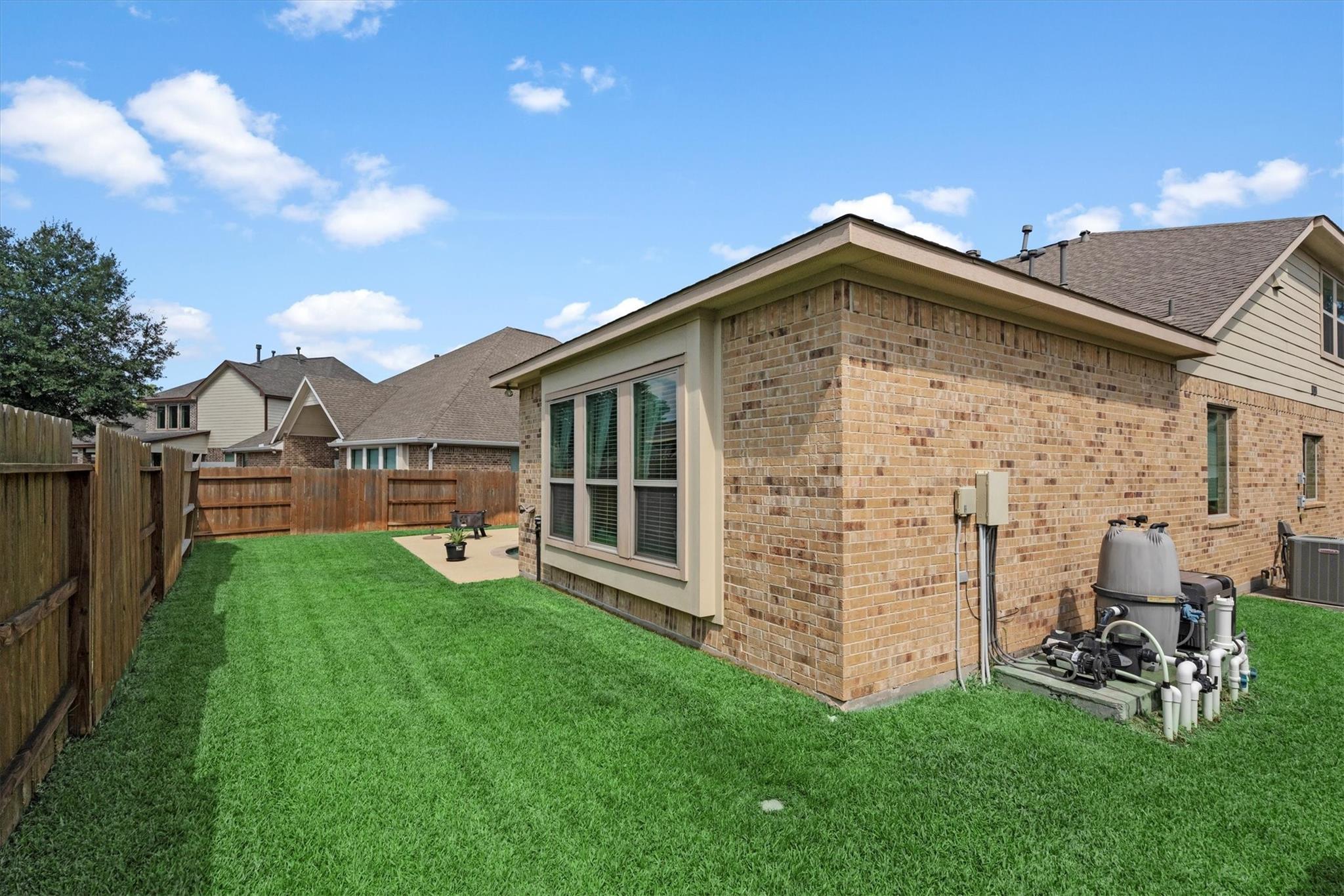 Fenced backyard with a wide lawn, small patio, and pool equipment beside a brick house.