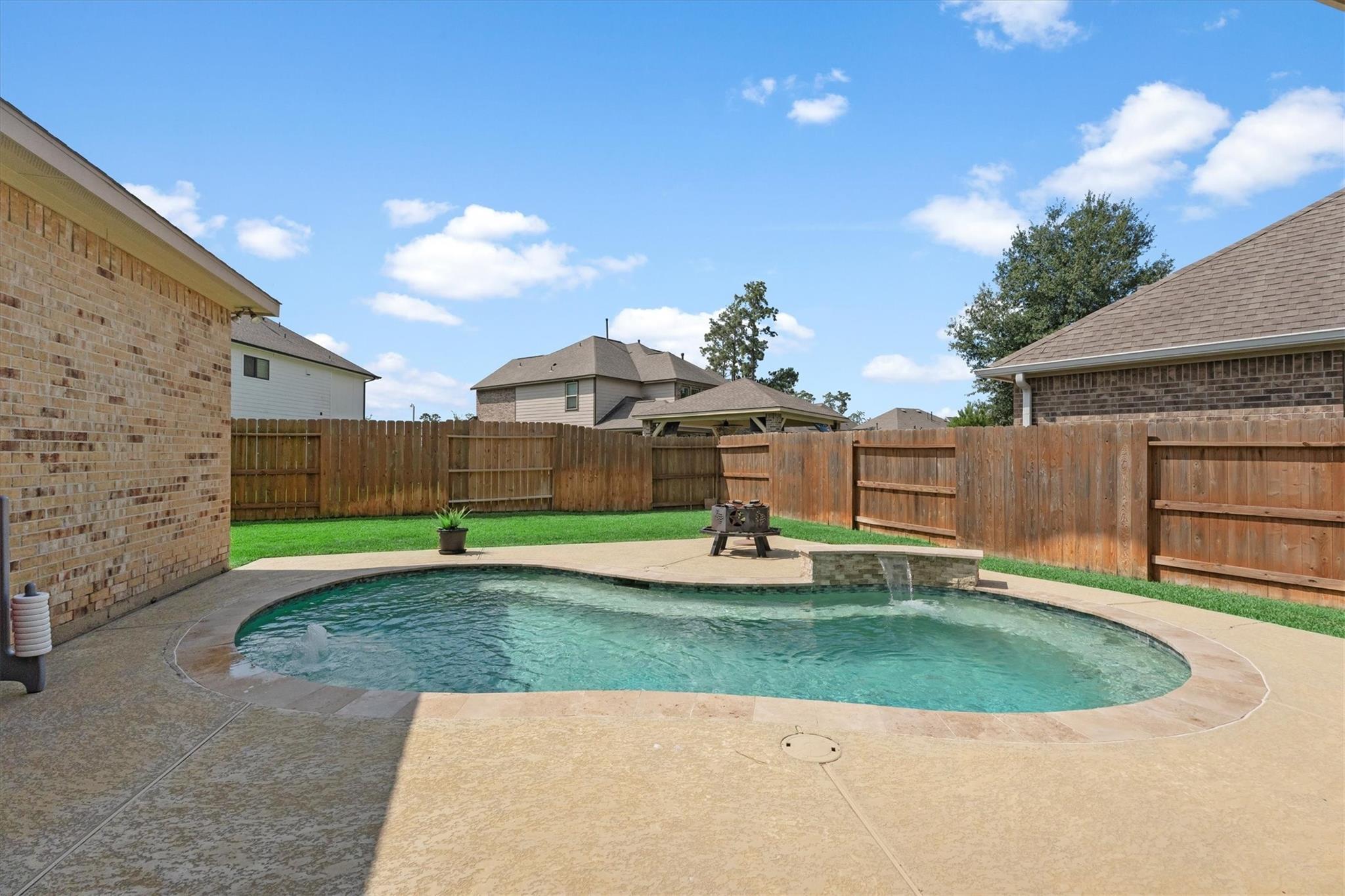 Backyard swimming pool with a small waterfall, concrete patio, lawn, and wooden privacy fence.