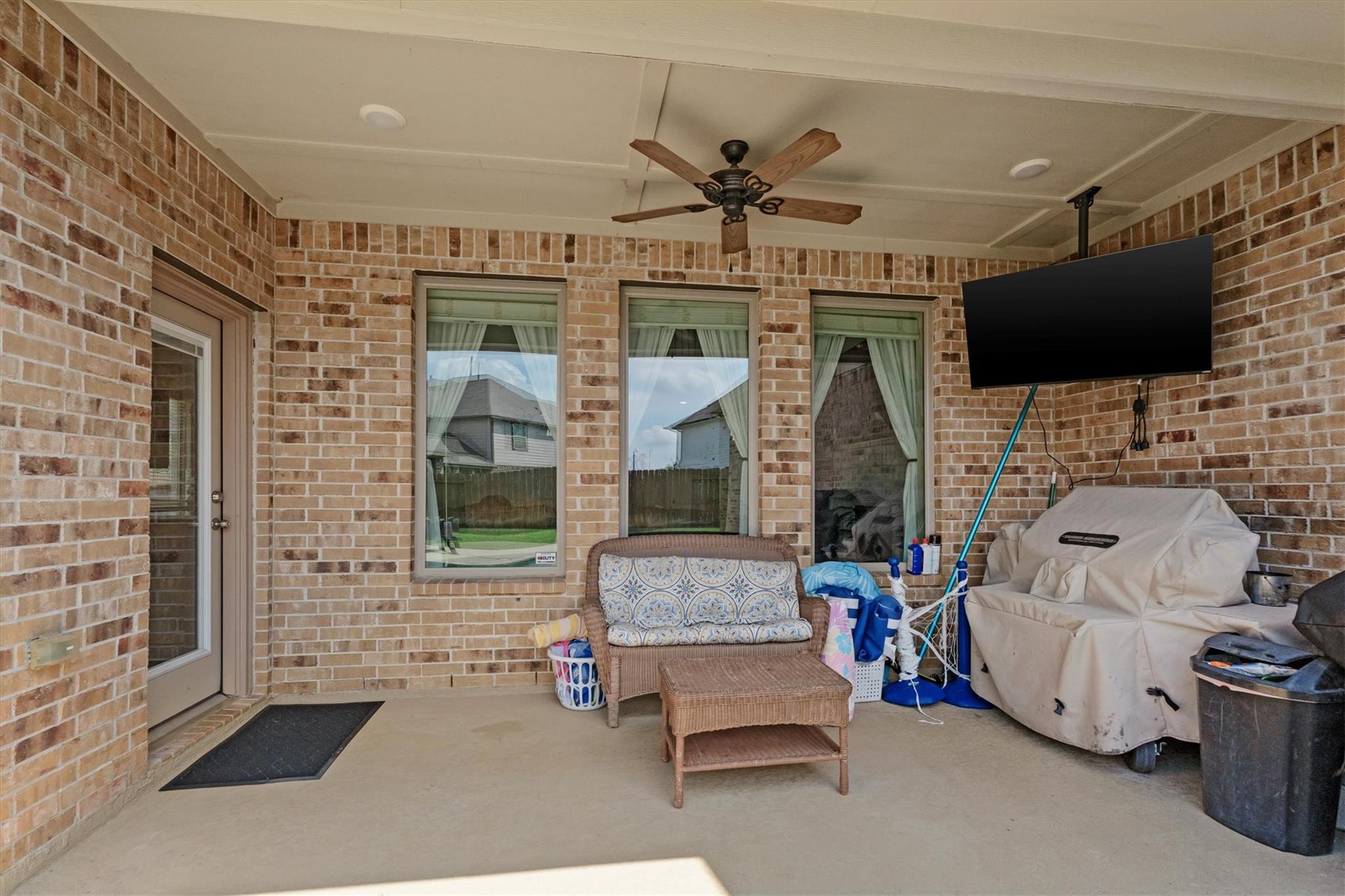 Covered brick patio with wicker seating, a ceiling fan, wall-mounted television, and covered grill.