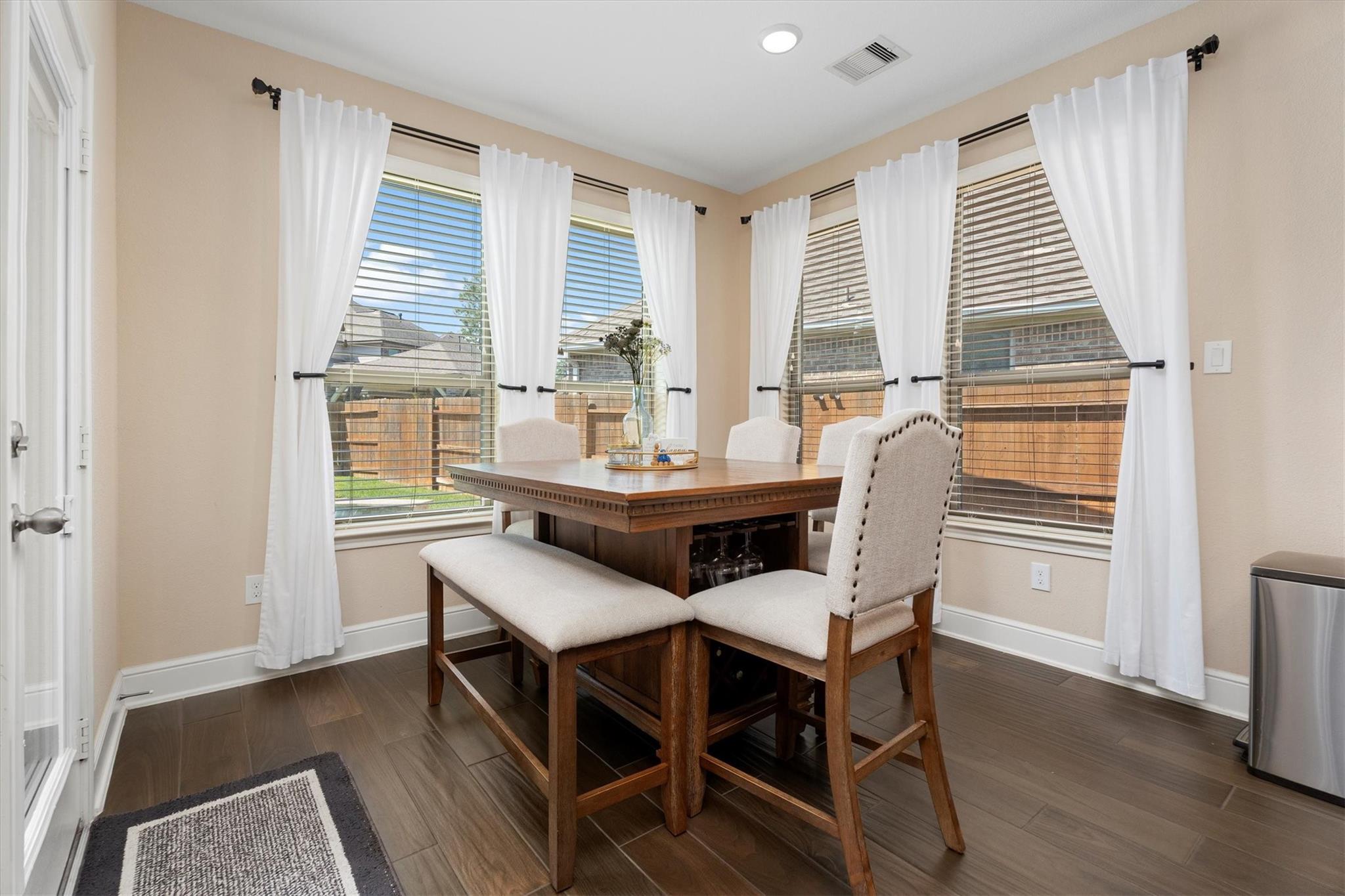 Dining room with a wooden table and upholstered seating beside windows with white curtains.