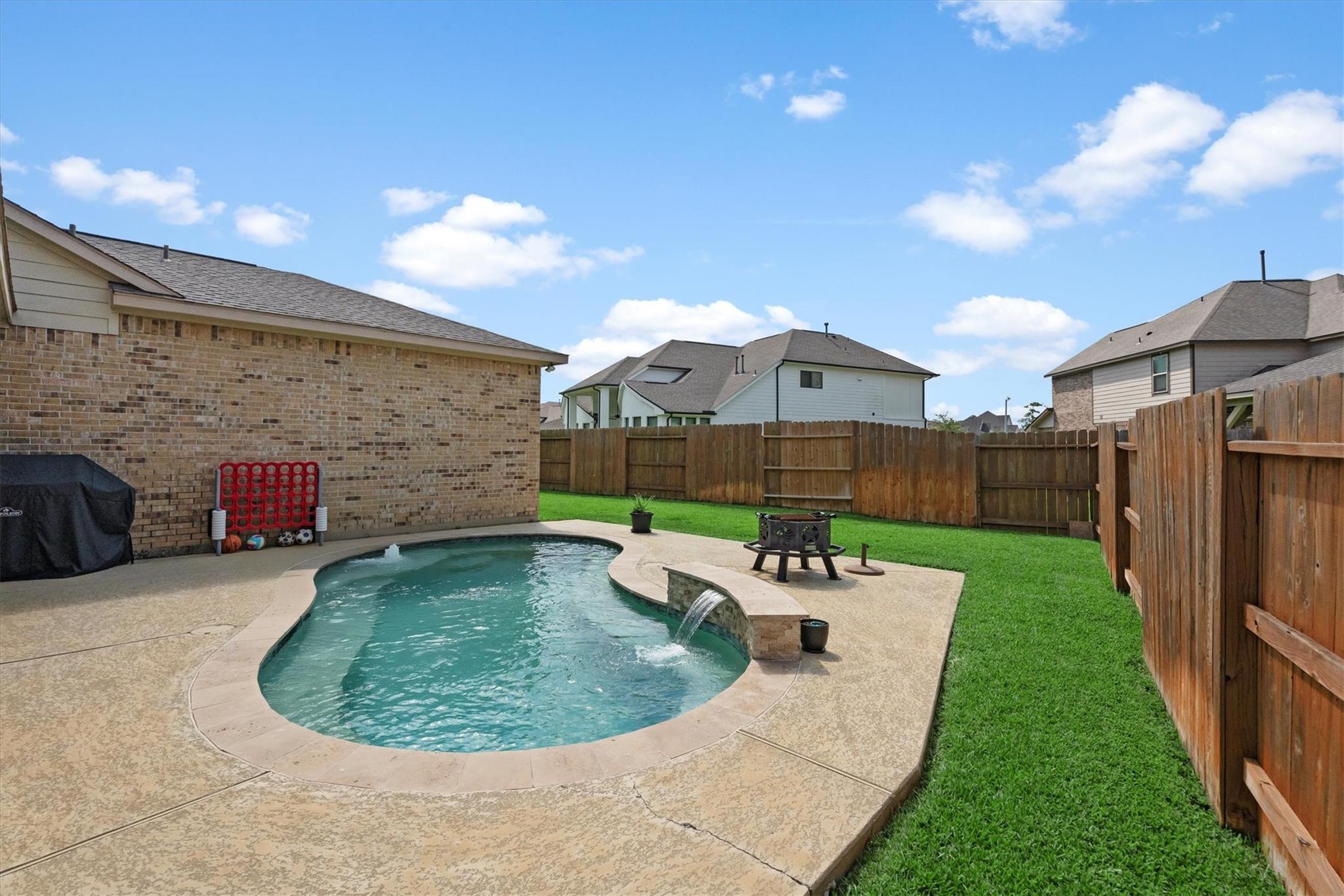 Curved backyard pool with a stone water feature, patio, lawn, and wooden privacy fence.