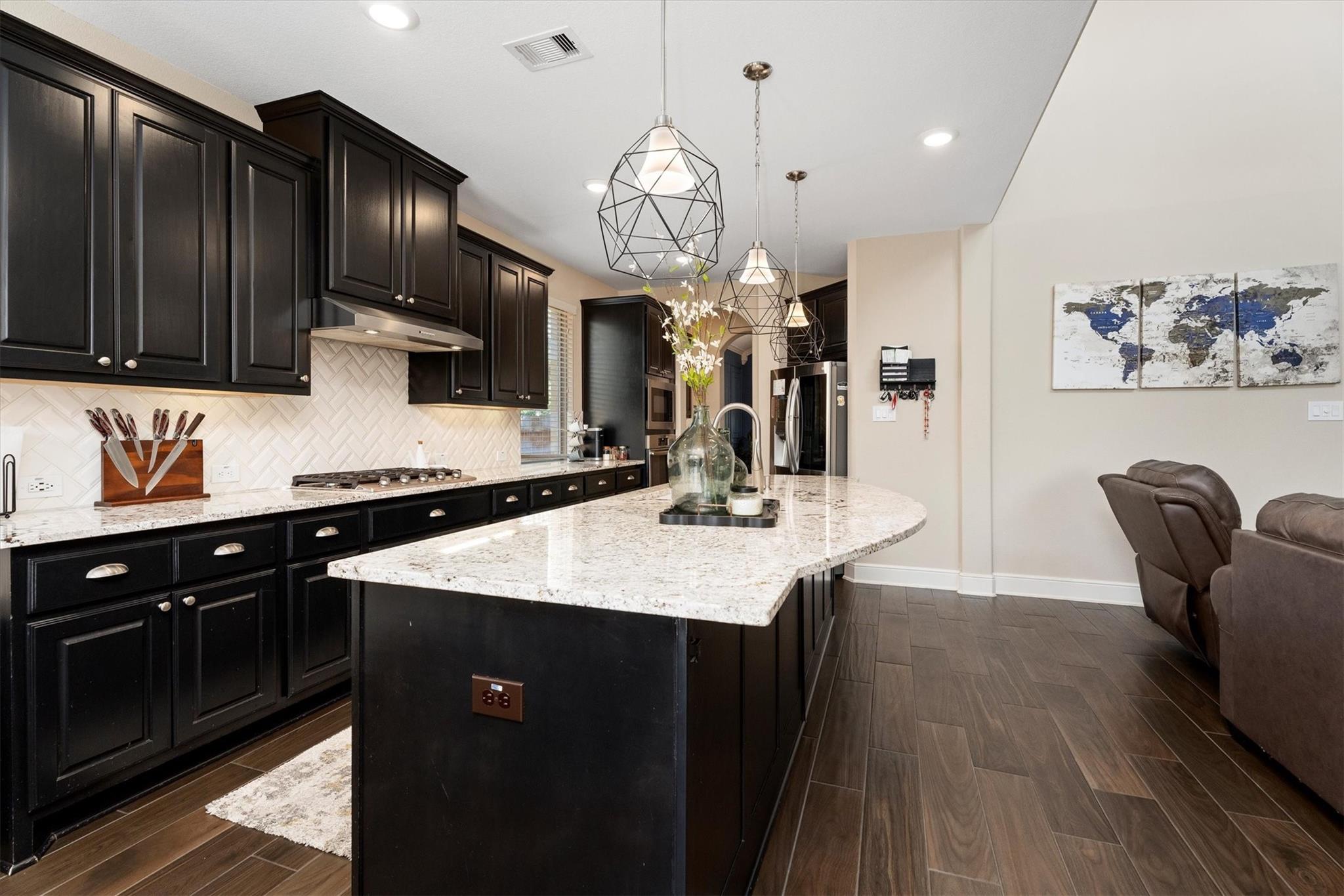 Kitchen with dark cabinets, a large granite island, geometric pendant lights, and an open view into the living area.