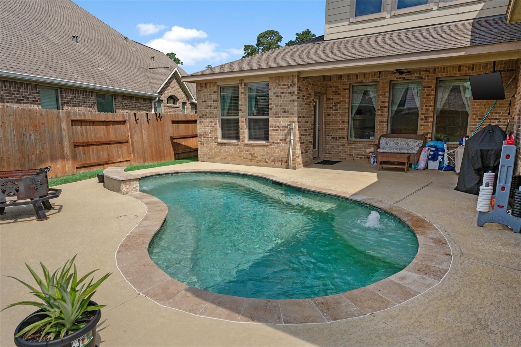 Curved backyard swimming pool beside a brick home, with a covered patio and fenced yard.