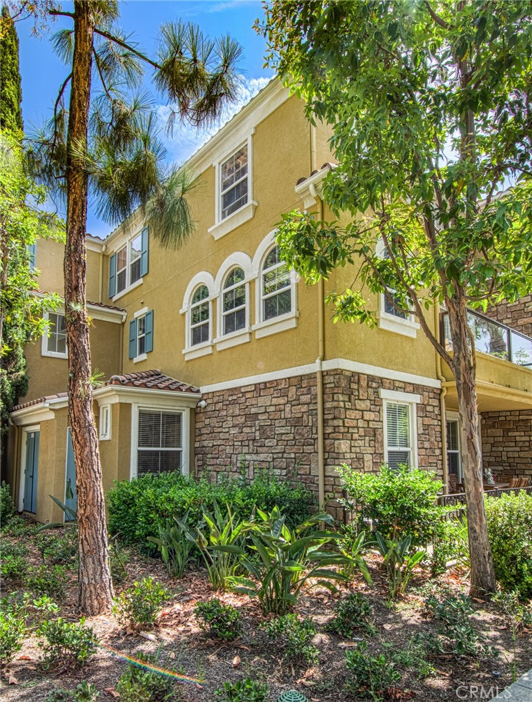 Two-story tan stucco and stone residence with arched windows, trees, and a landscaped front yard.