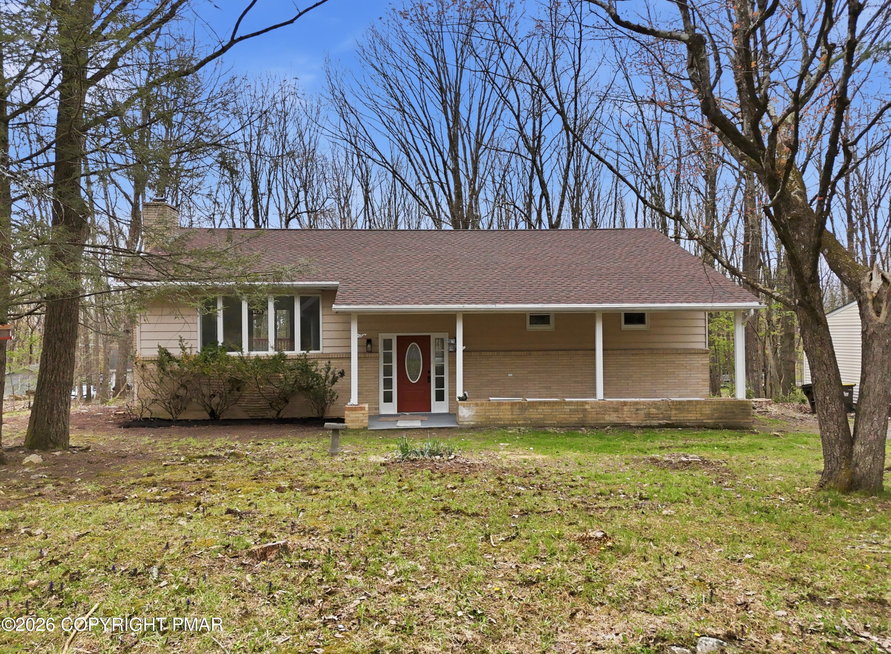 Tan brick-and-siding ranch home with a long covered porch and red front door among leafless trees.