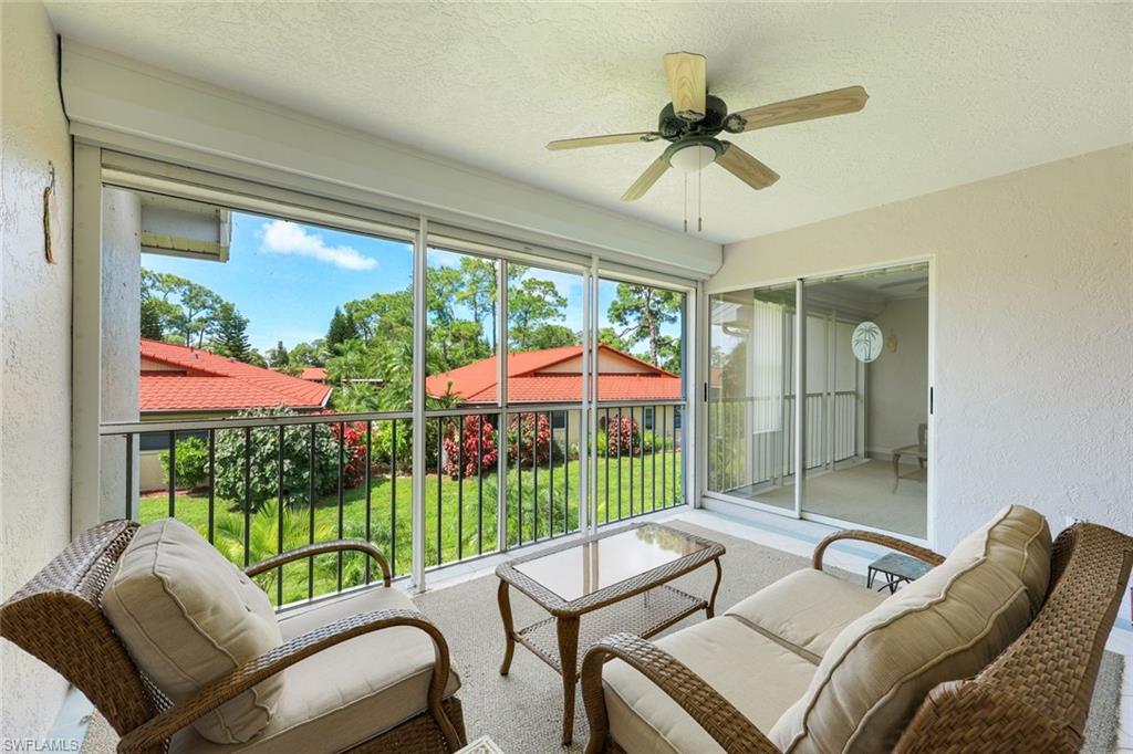 Furnished enclosed lanai with wicker seating and views of a lawn, flowering shrubs, trees, and red-tile roofs.