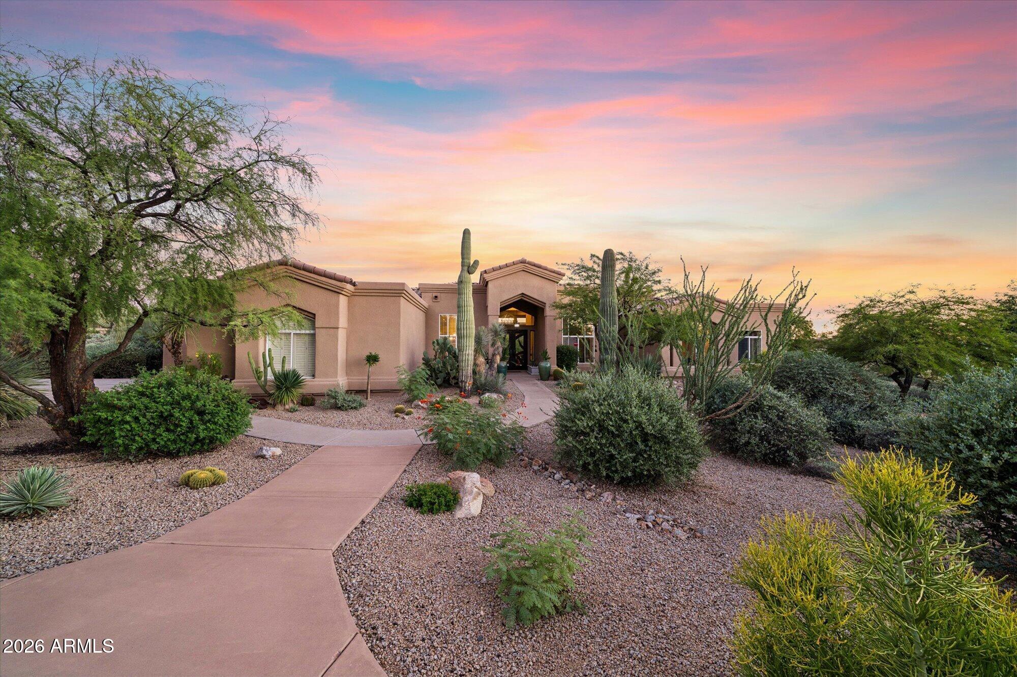 Stucco home with a curved walkway and desert landscaping beneath a colorful dusk sky.