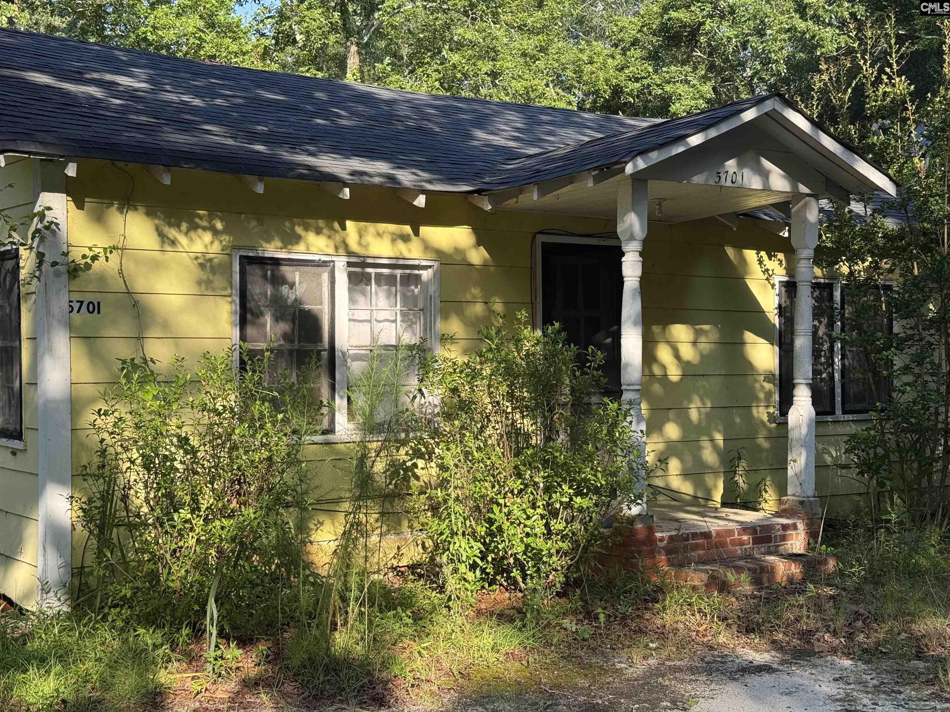 Yellow house with a covered porch, brick steps, and overgrown shrubs.